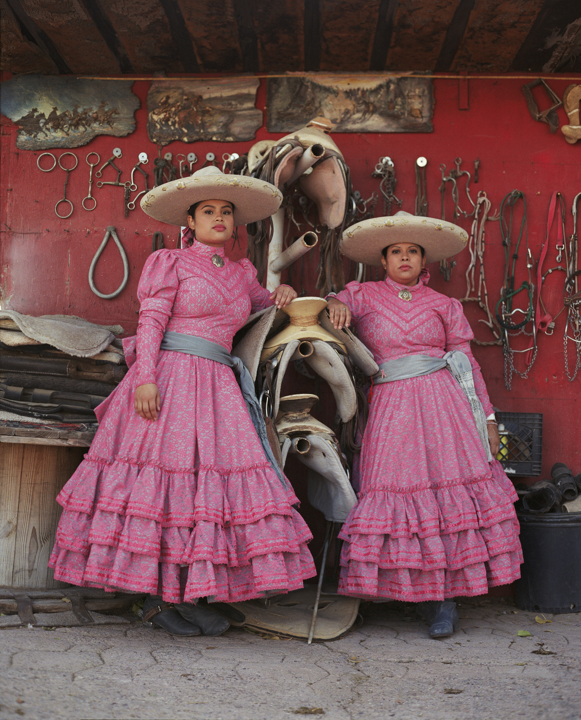 Two women in pink ruffled dresses and white sombreros pose beside horse sculptures against red wall displaying bridles and equestrian equipment.