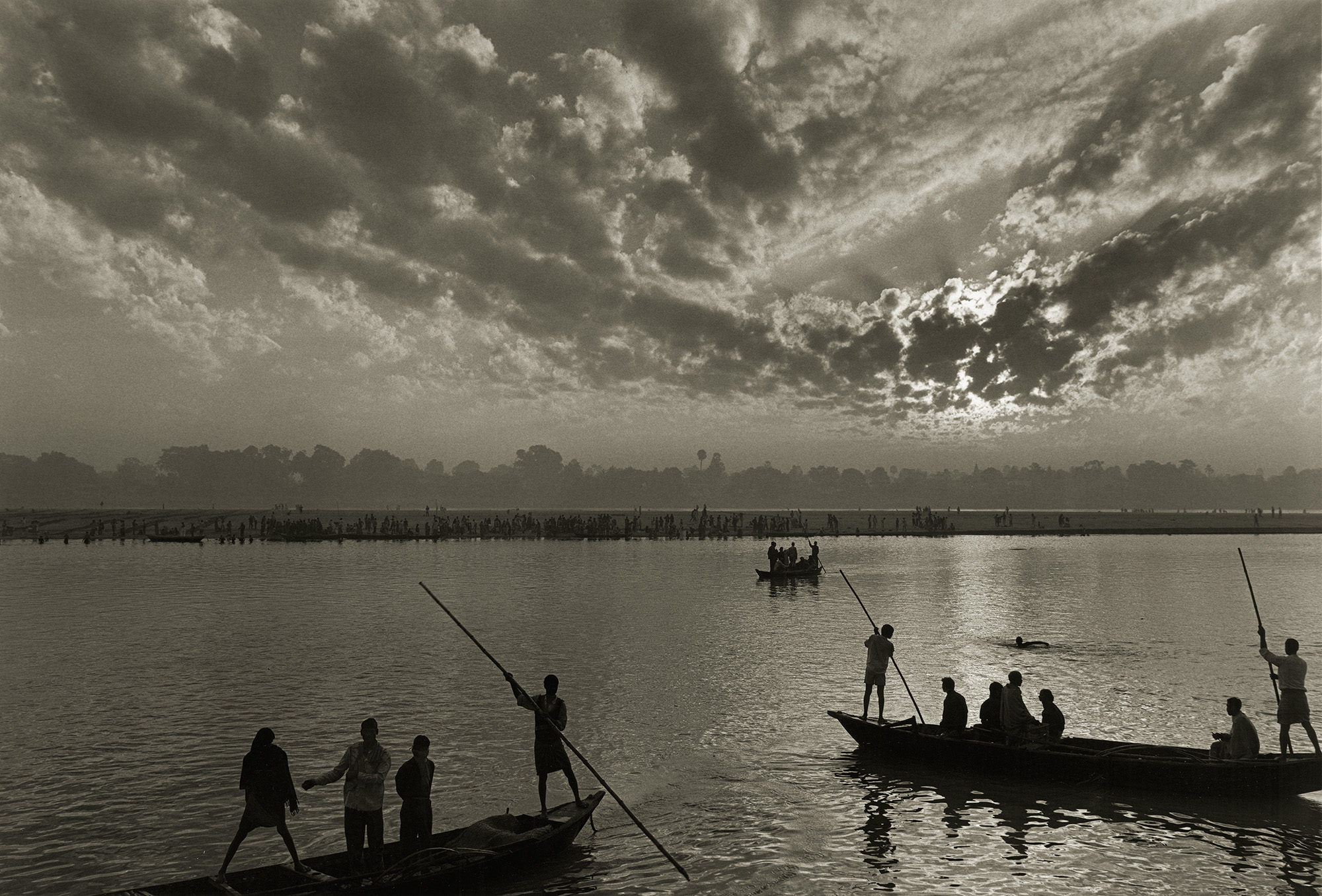 Silhouetted figures with long poles on boats in calm water under dramatic cloudy sky. Monochrome image with strong contrast.