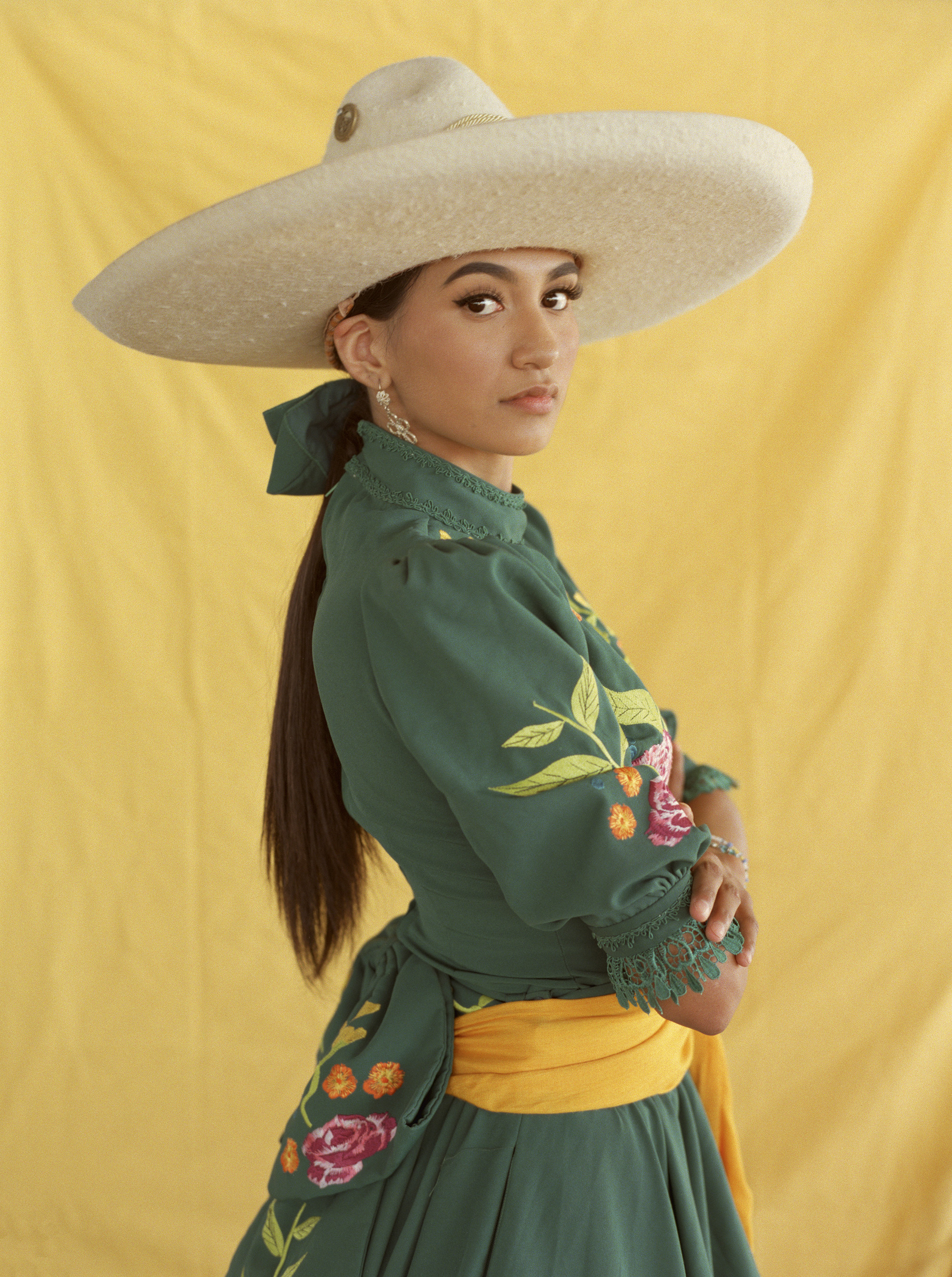 Woman in wide-brimmed white hat and green dress with floral embroidery, yellow sash at waist, against yellow background.