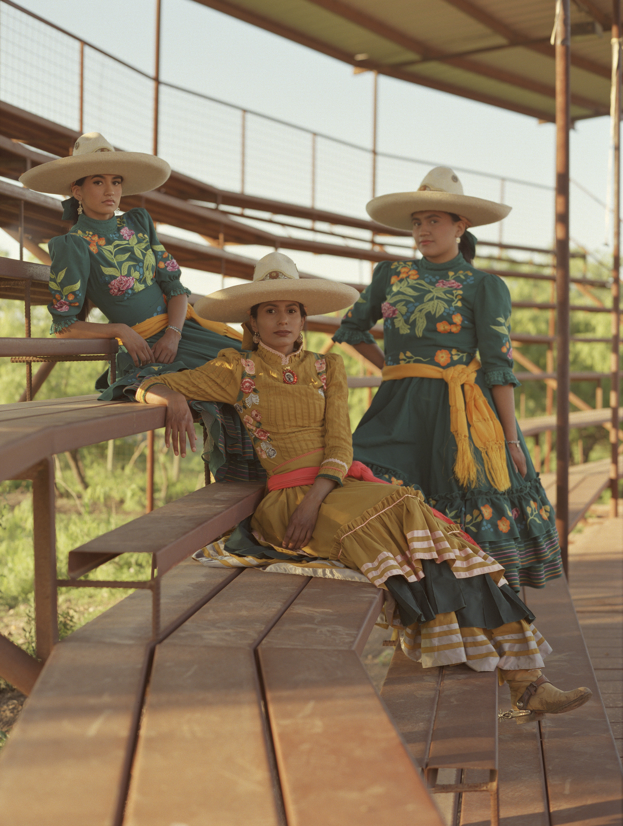 Three women in colourful traditional Mexican dresses and wide-brimmed hats sitting on wooden bleacher steps with metal railings.