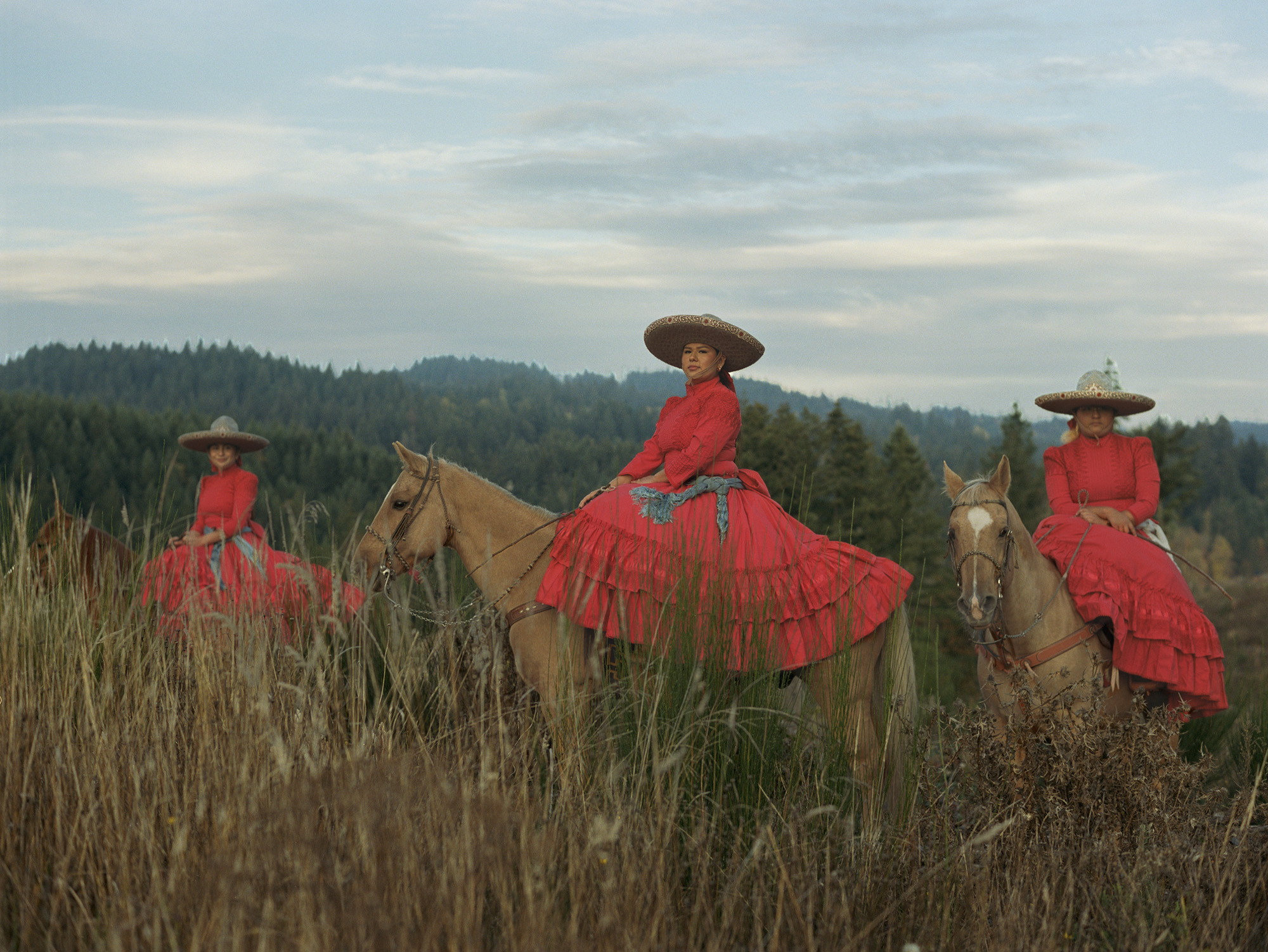 Three riders in bright red dresses and wide-brimmed hats on horses in tall grass field with forested hills and cloudy sky behind.