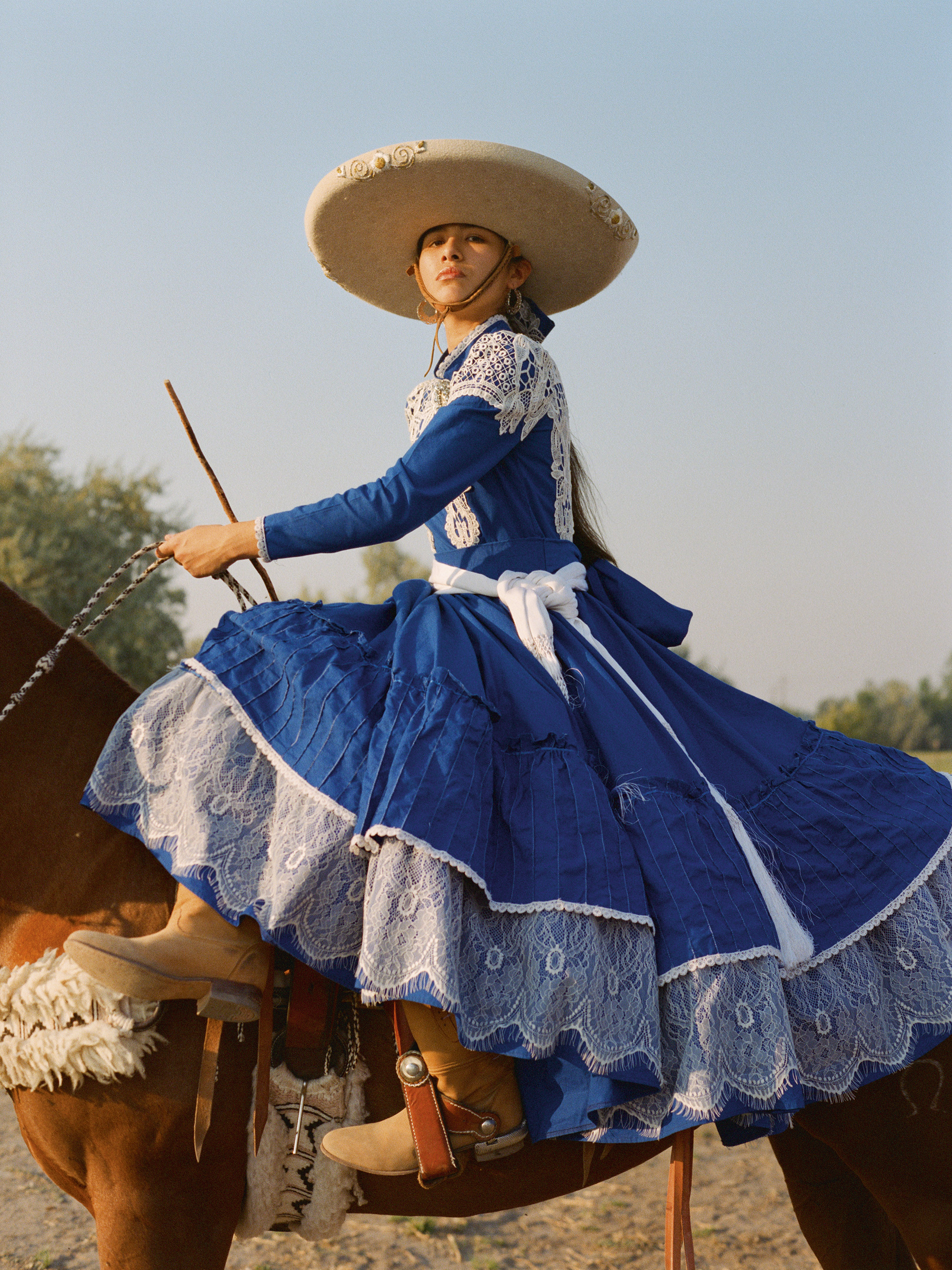 Woman in blue and white Mexican traditional dress with large sombrero riding horse, holding reins against cloudy sky.