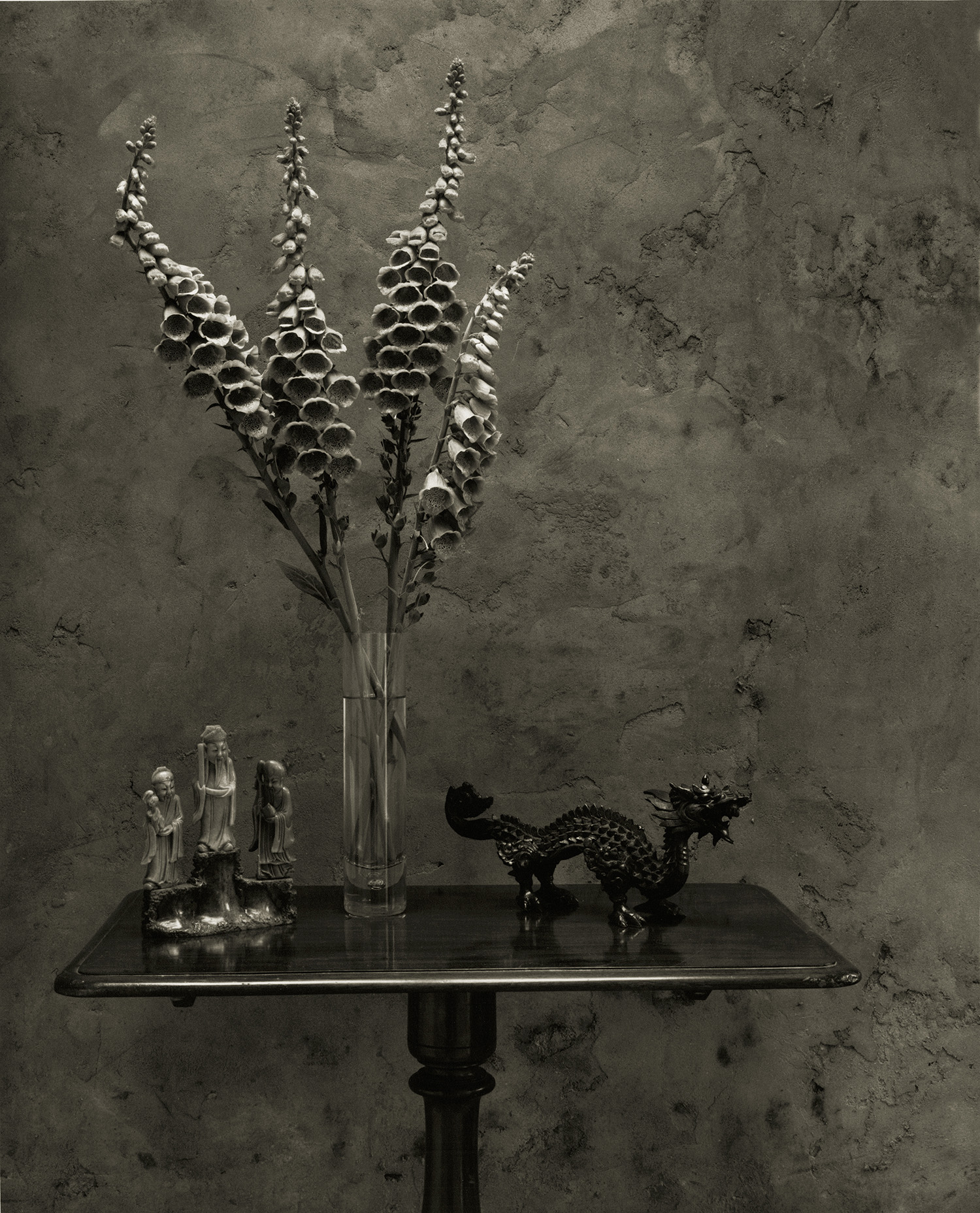 Clear glass vase with tall dried flower stems on dark green table, alongside small decorative figurines and ornate metal objects.