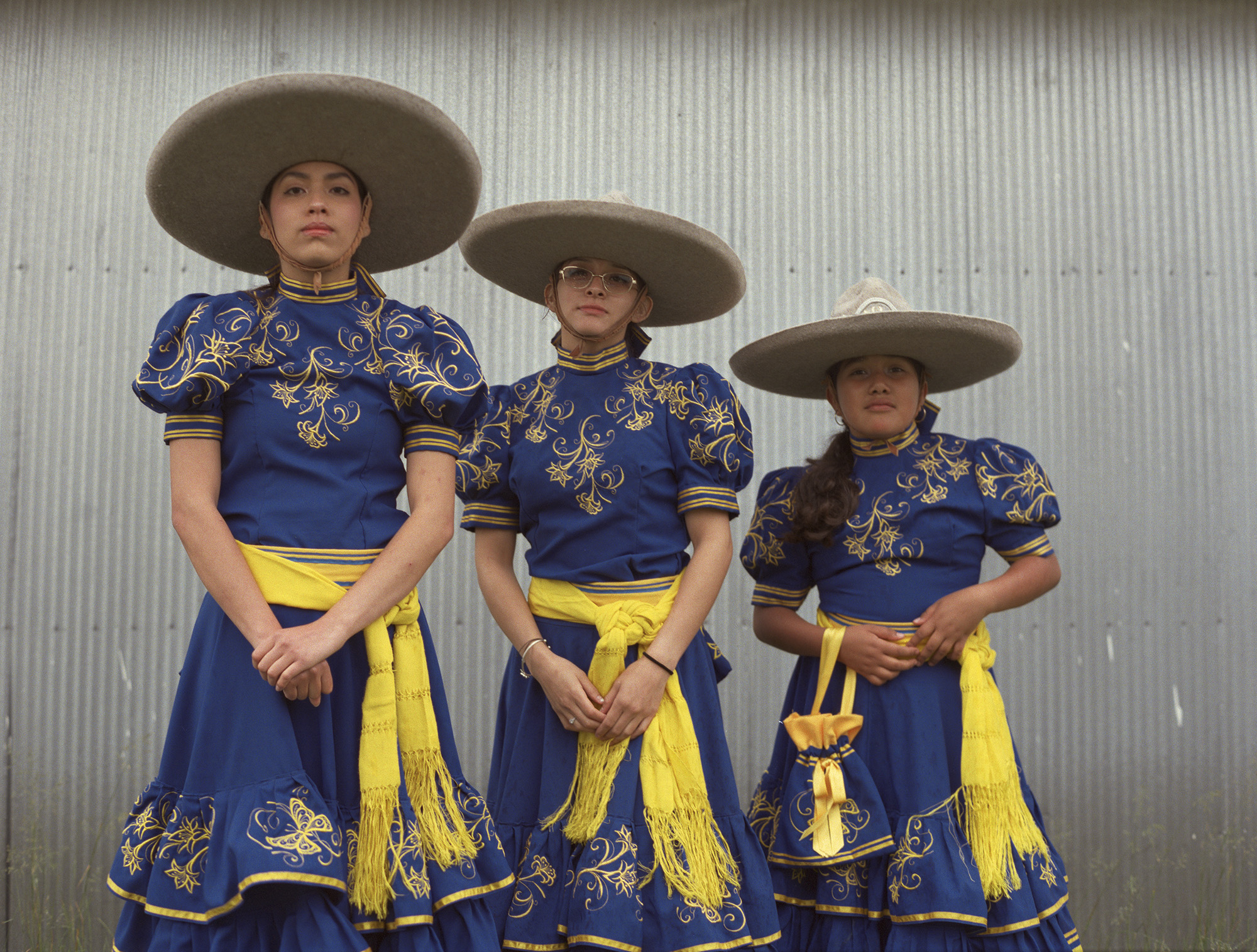 Three women in traditional Mexican charro outfits with blue embroidered dresses, yellow sashes, and wide-brimmed hats against metal wall.
