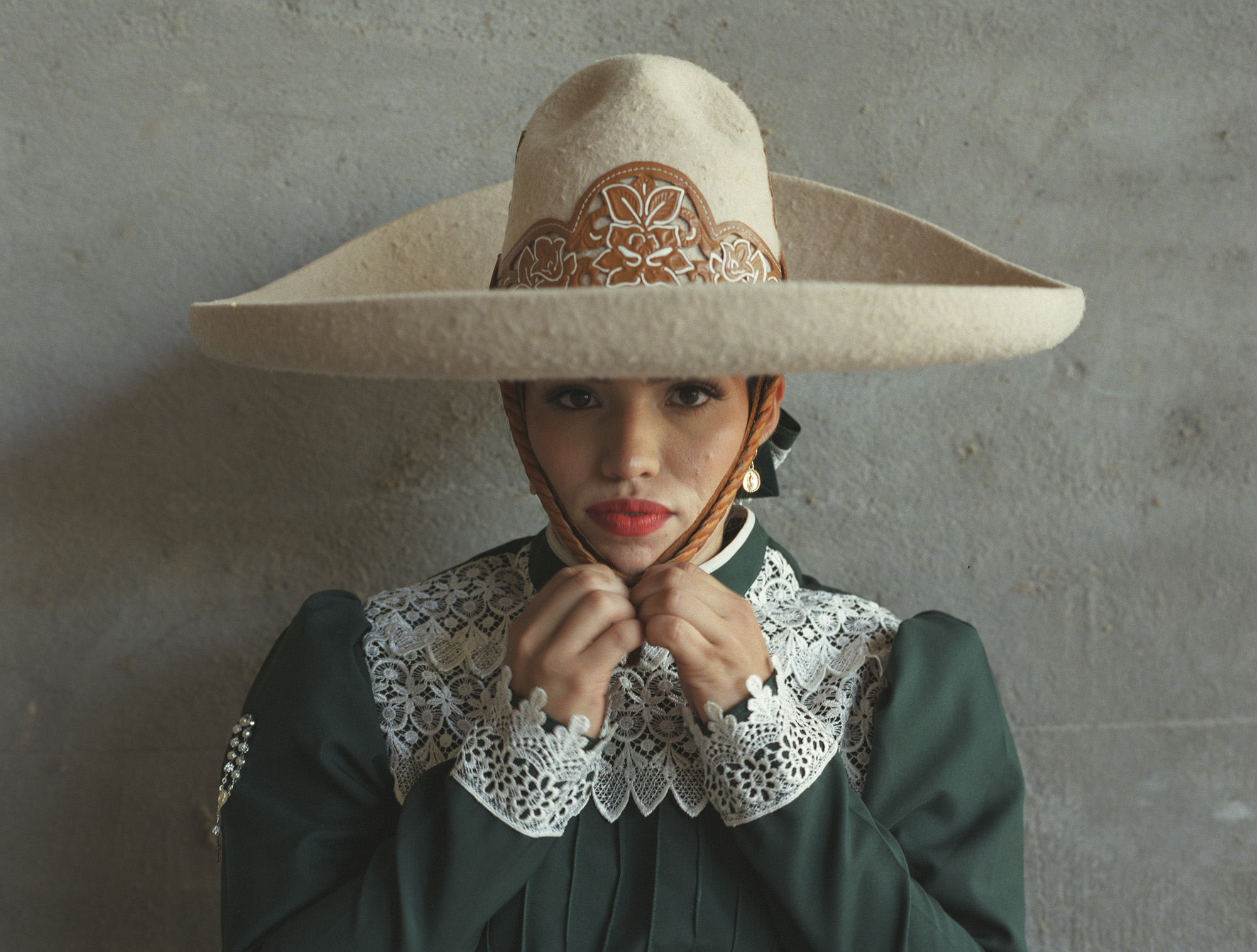 Woman wearing wide-brimmed cream hat with decorative band and green dress with white lace collar against grey textured wall.
