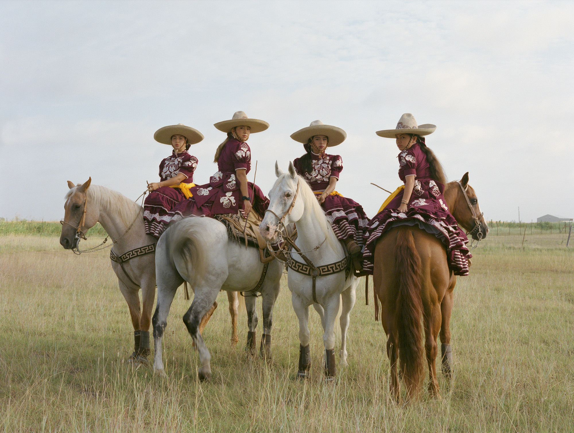 Four riders in wide-brimmed hats and colourful traditional clothing mounted on horses in an open grassland field under cloudy skies.