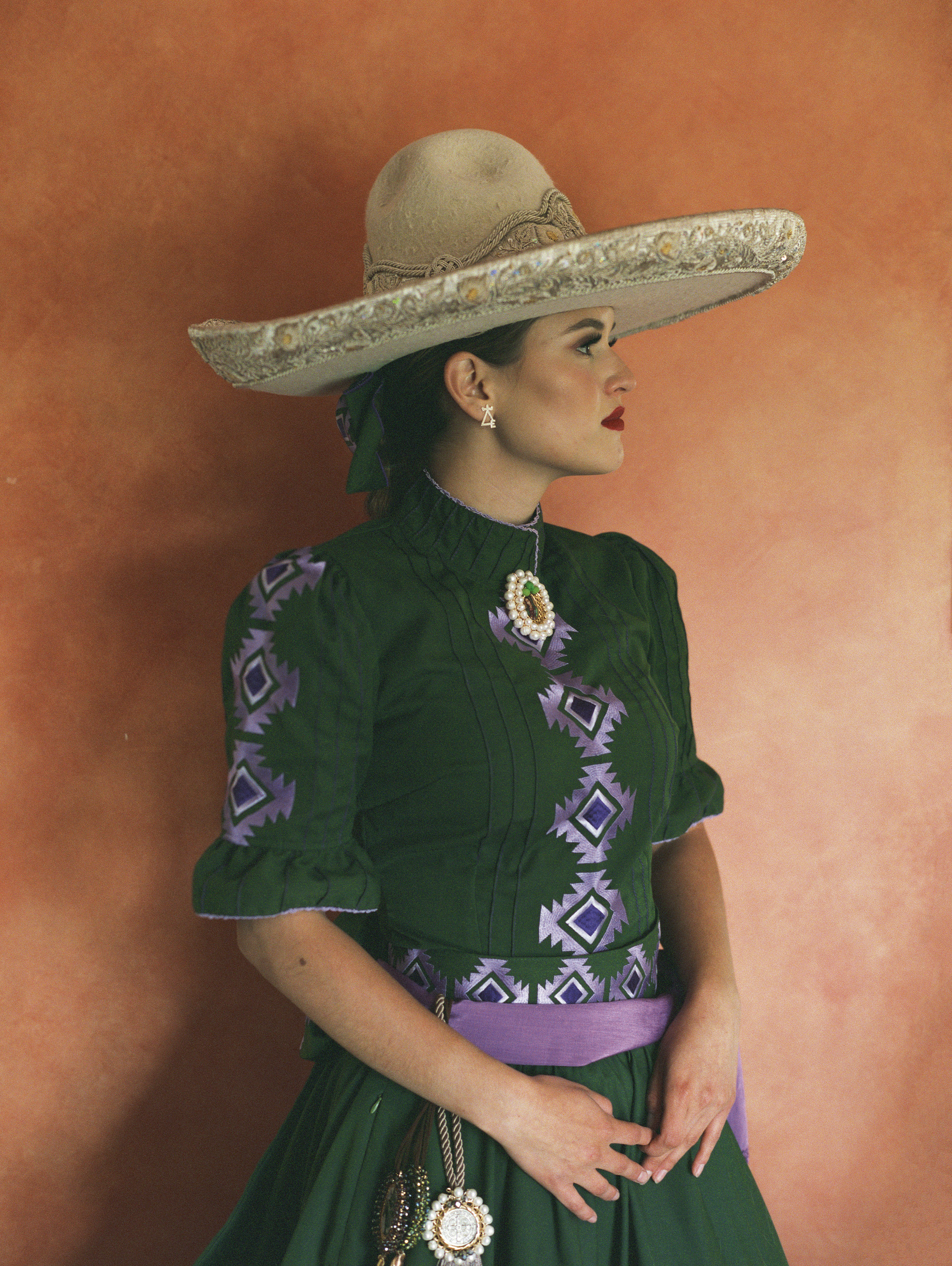 Woman in profile wearing wide-brimmed straw hat and green dress with purple geometric patterns against orange wall background.