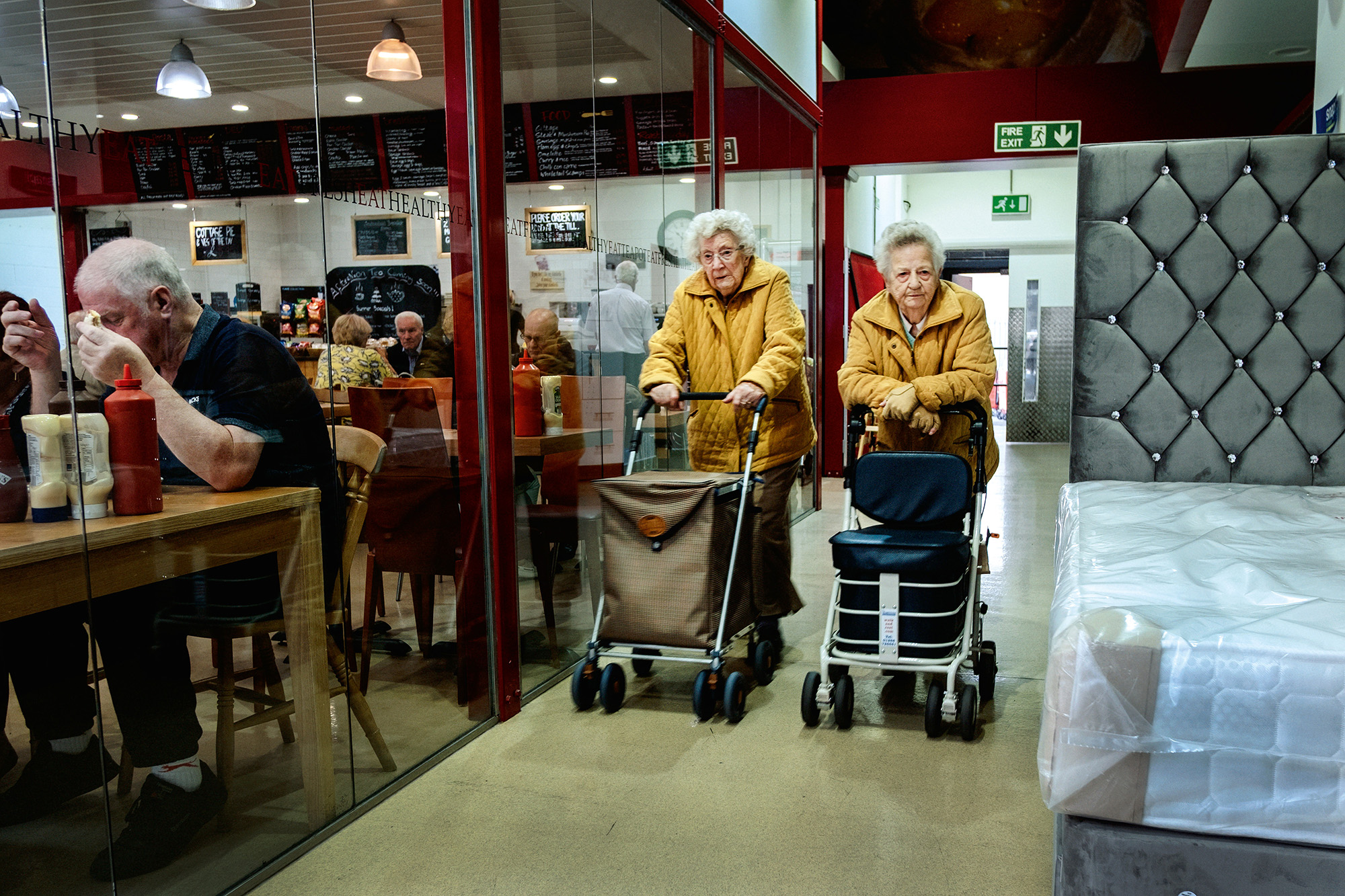 Two elderly women with walking frames stand behind glass partition separating them from seated diners in restaurant or café setting.