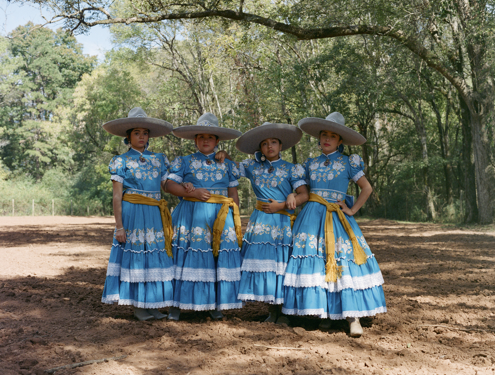 Four women in matching blue traditional dresses with white floral patterns and yellow sashes, wearing wide-brimmed hats, standing outdoors.