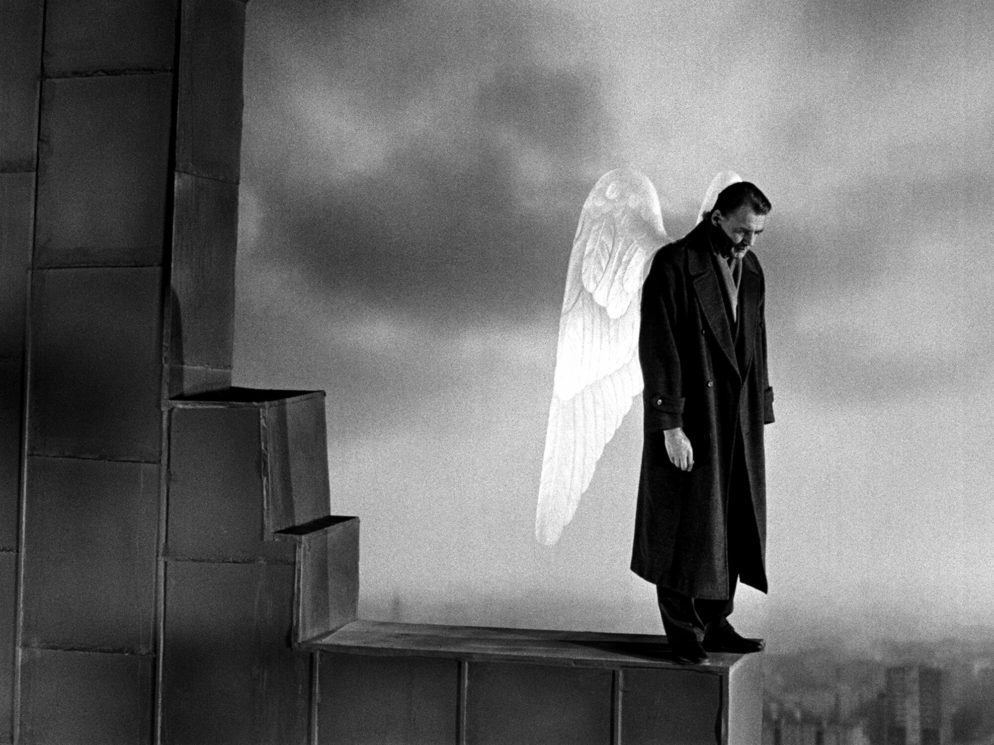 Lone figure in long dark coat stands atop steps, silhouetted against bright white wings and cloudy sky.