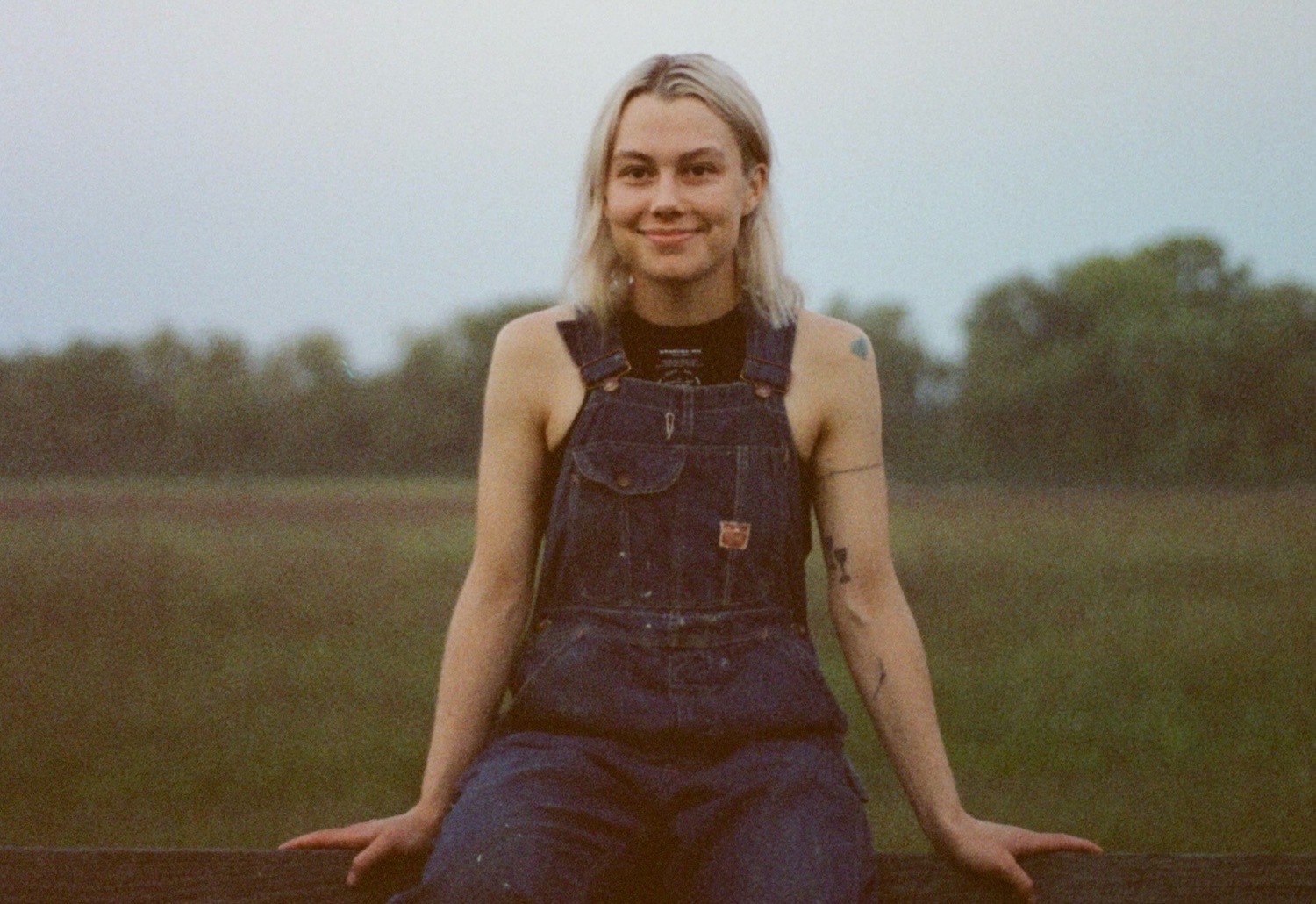 Phoebe Bridgers sitting on fence dungarees
