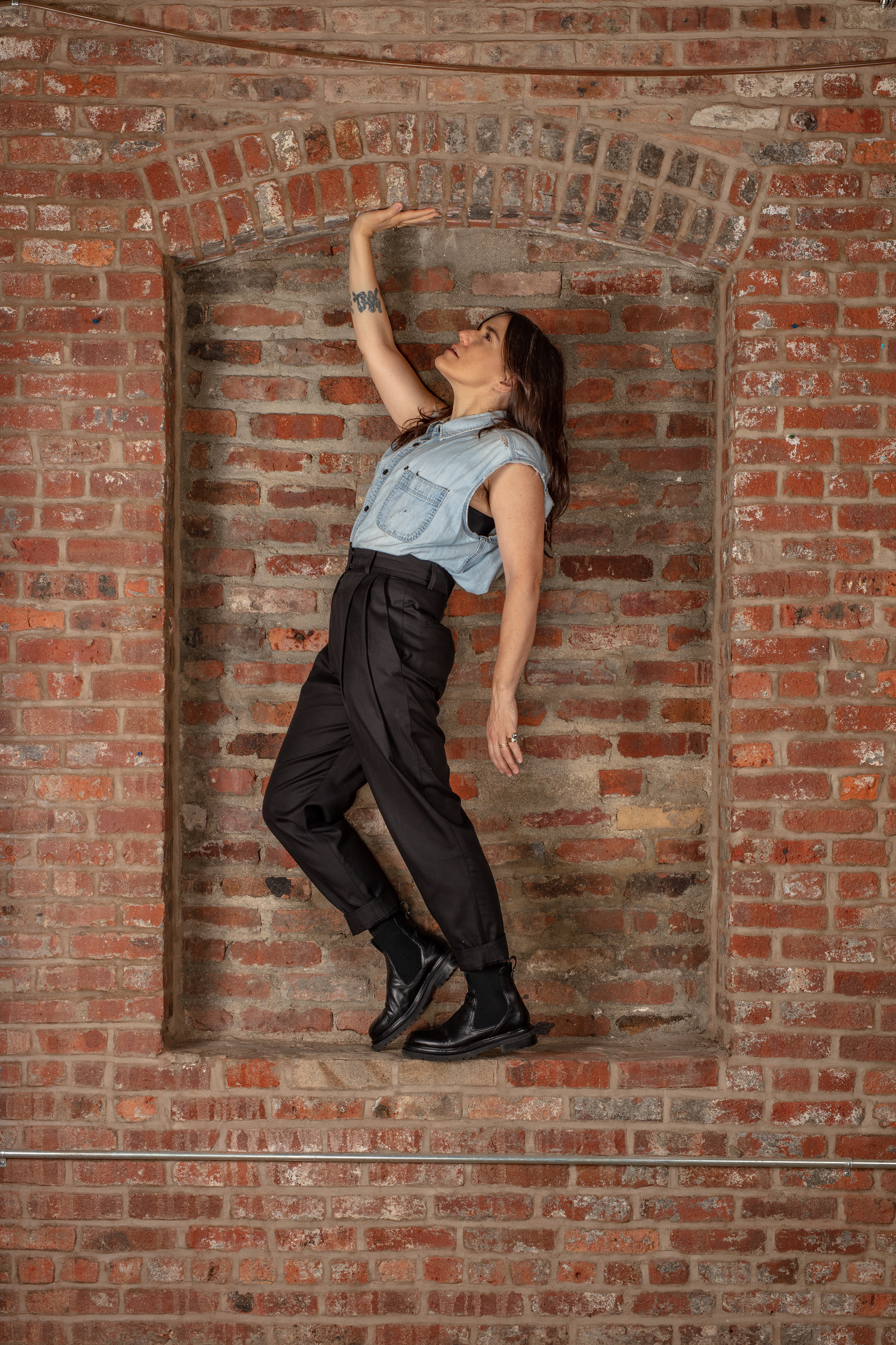 Emily Wells in black trousers and a blue top standing in an alcove of a brick wall