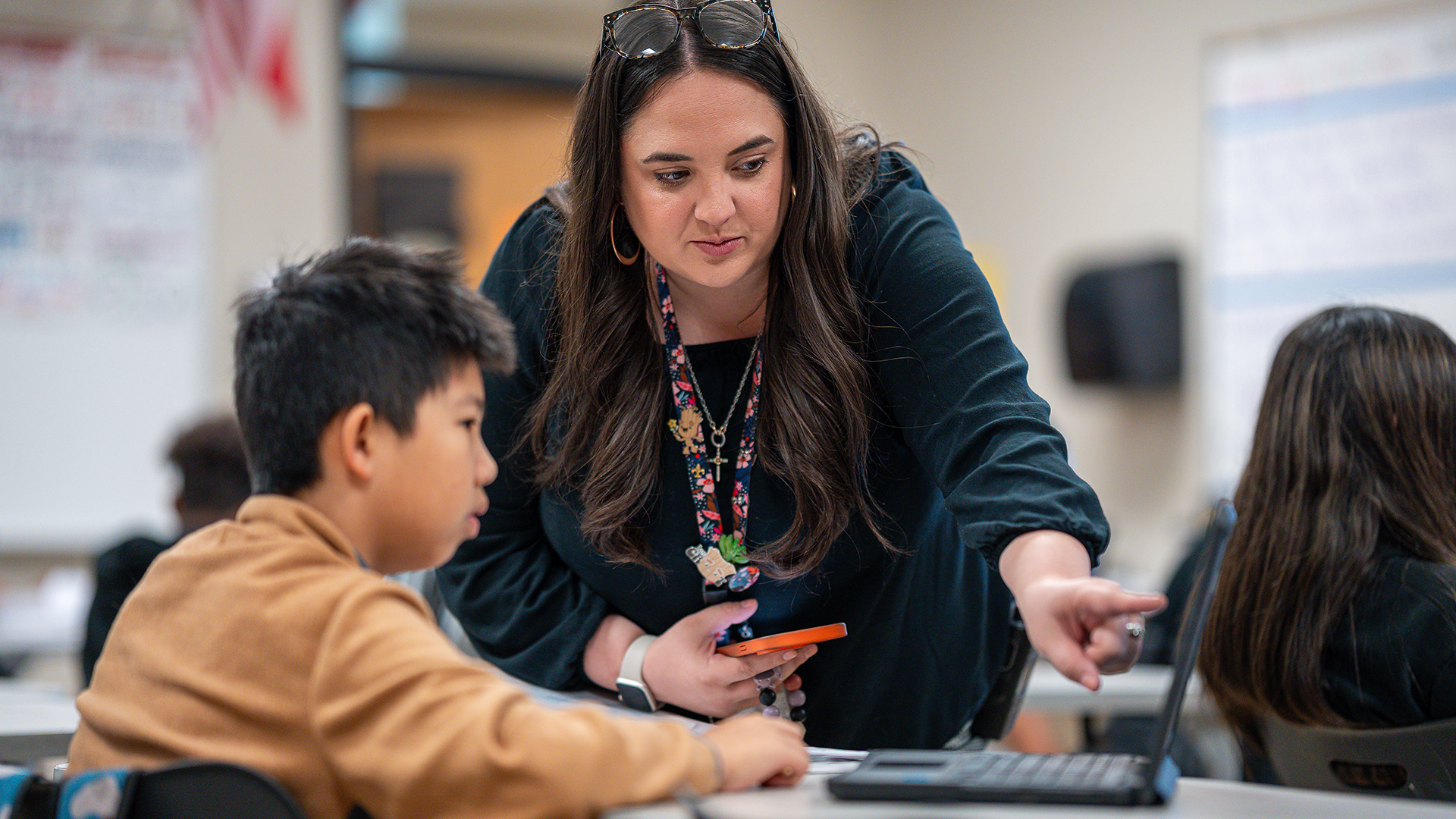Teacher leans over a student, pointing at a laptop while holding an orange phone and wearing a floral lanyard and glasses on her head.
