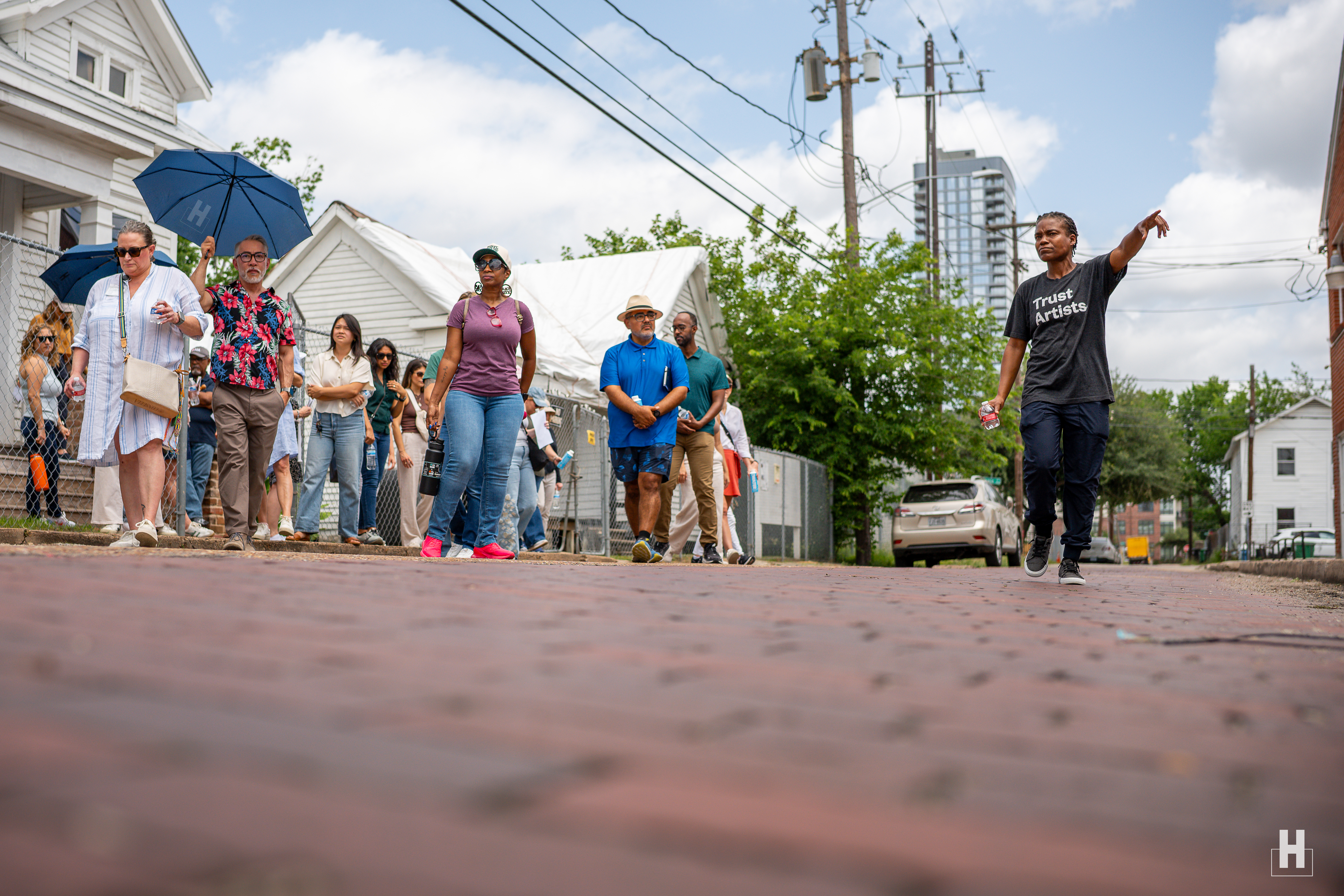 Group of people walking on a brick street past white houses and a fence, one woman in a 'Trust Artists' shirt pointing ahead.