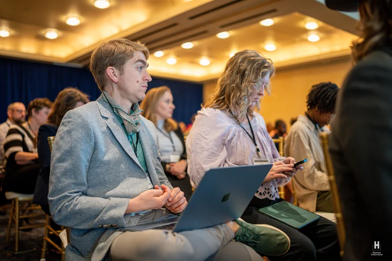 Attentive attendees seated in a warm ballroom: man in gray blazer with laptop and green scarf beside a woman scrolling her phone.