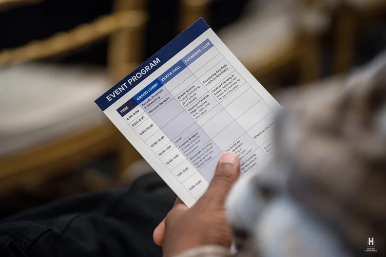 Hand holding a folded event program schedule card with navy header and timetable columns, thumb visible, blurred chairs in background.