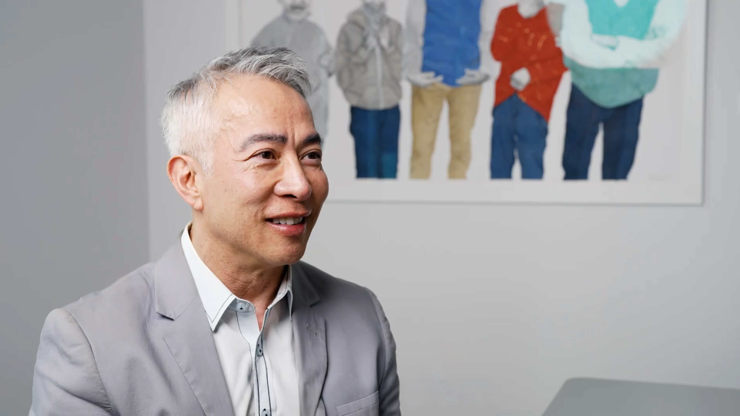 Smiling middle-aged person with short gray hair in a light-gray blazer, seated at a table, colorful framed print of four figures behind.