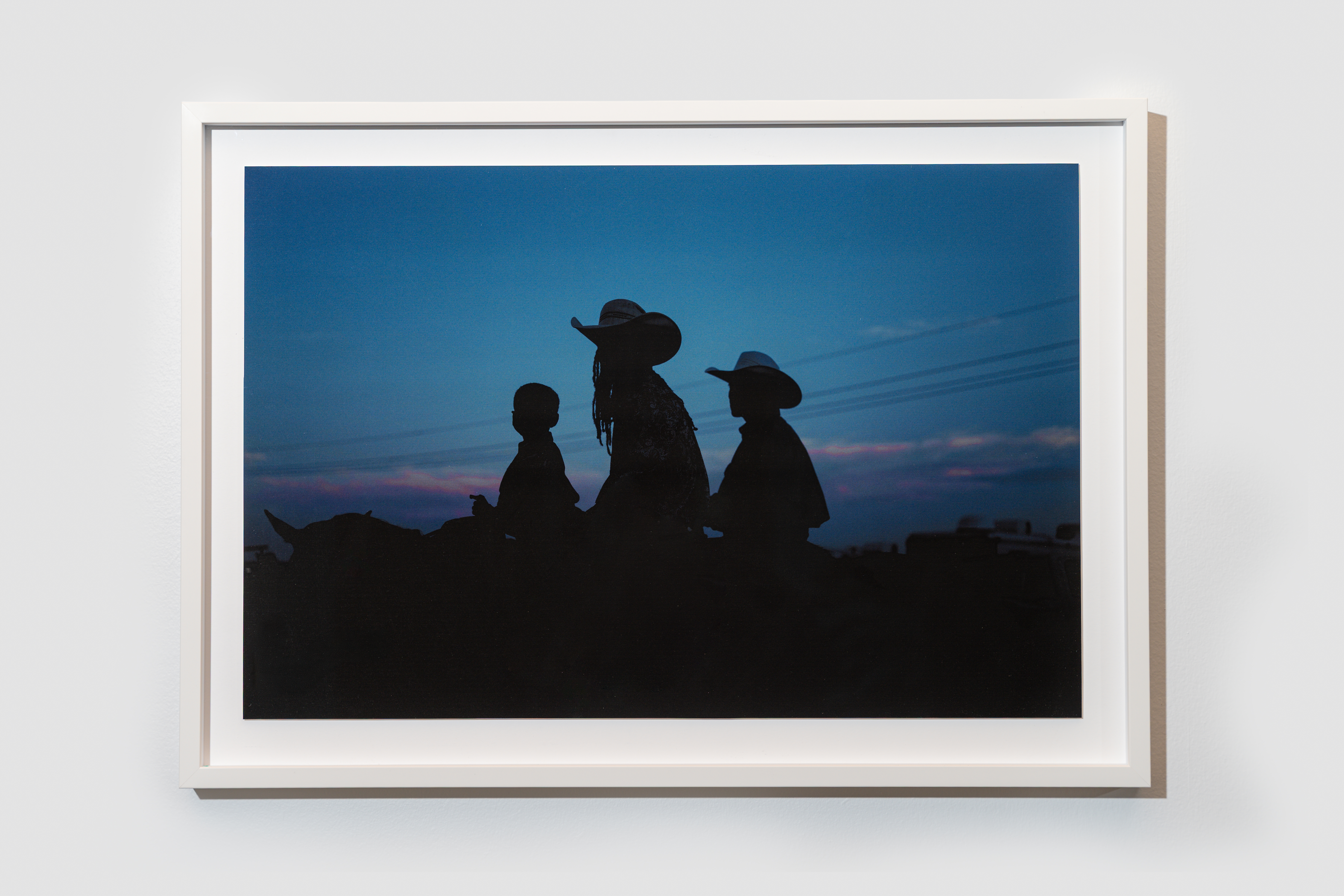 Silhouetted trio of horseback riders wearing cowboy hats against a deep blue twilight sky with pink horizon, framed on a white wall.