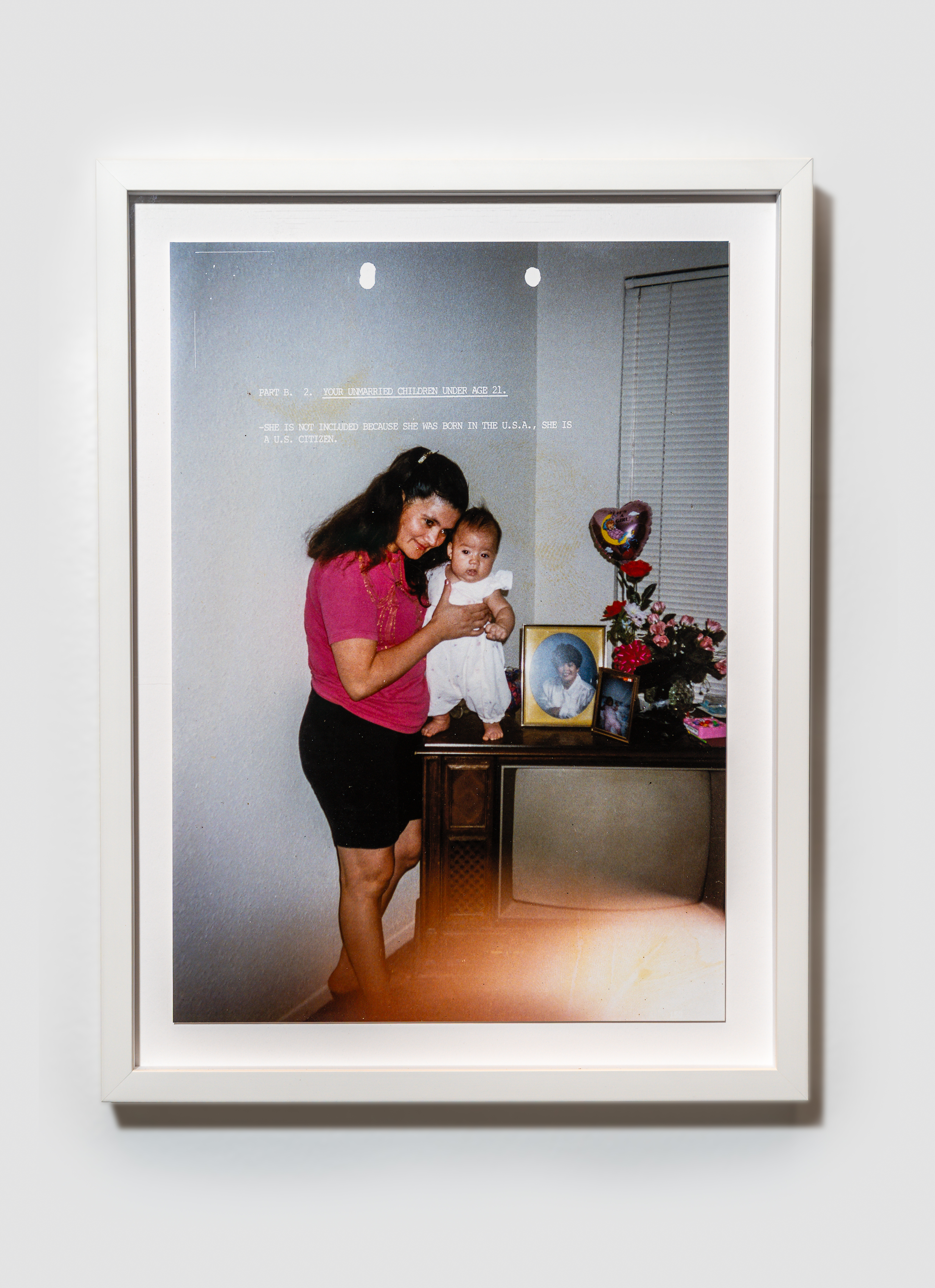 White-framed photograph shows a woman in pink holding a baby on a TV console beside framed photos, flowers and a heart-shaped balloon.
