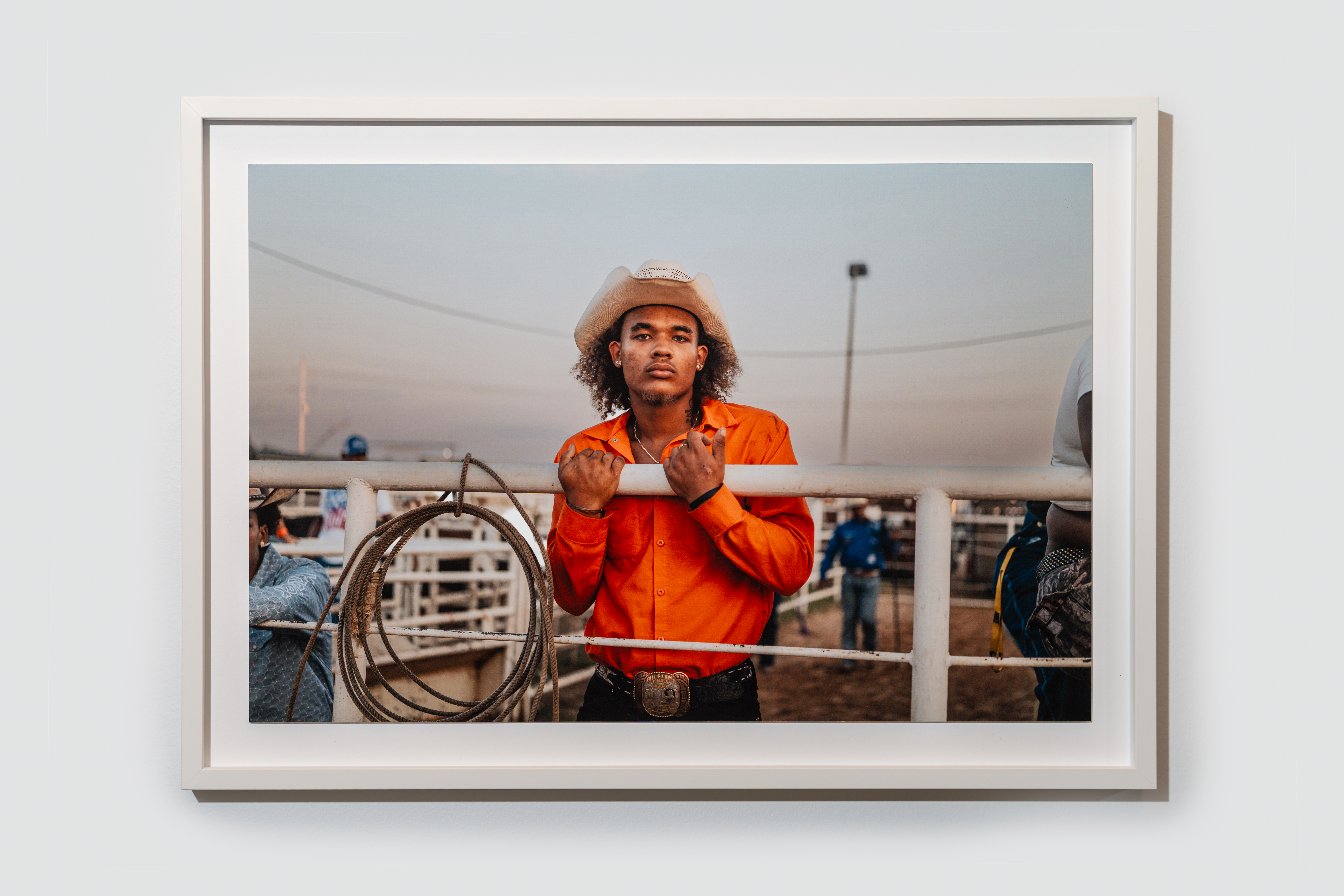 Young cowboy in a white hat and orange shirt grips metal fence at a rodeo pen, coiled lariat beside him at sunset, belt buckle visible.