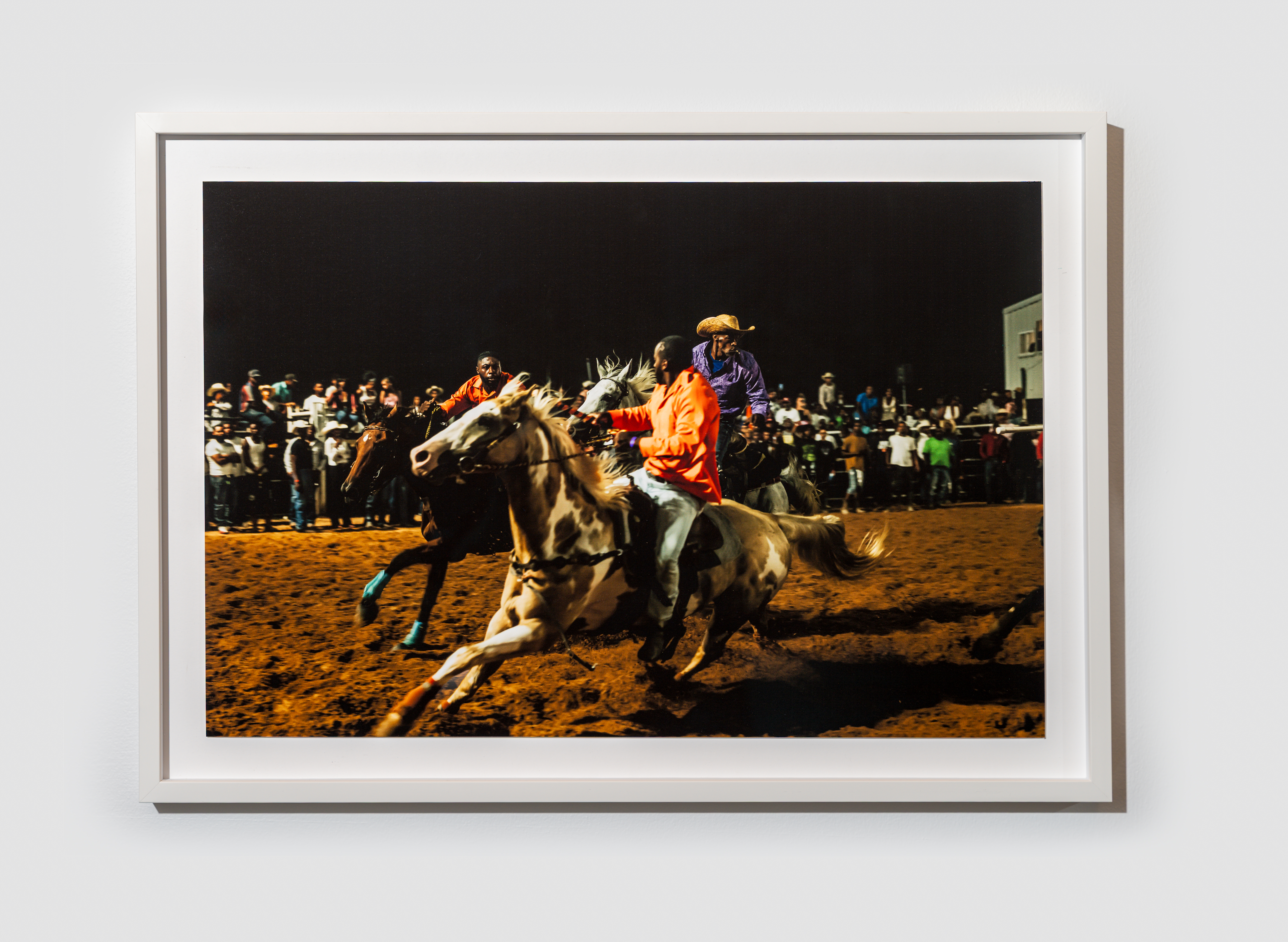 Night rodeo scene with two riders in bright orange and purple jackets racing horses across muddy arena as a cheering crowd watches.