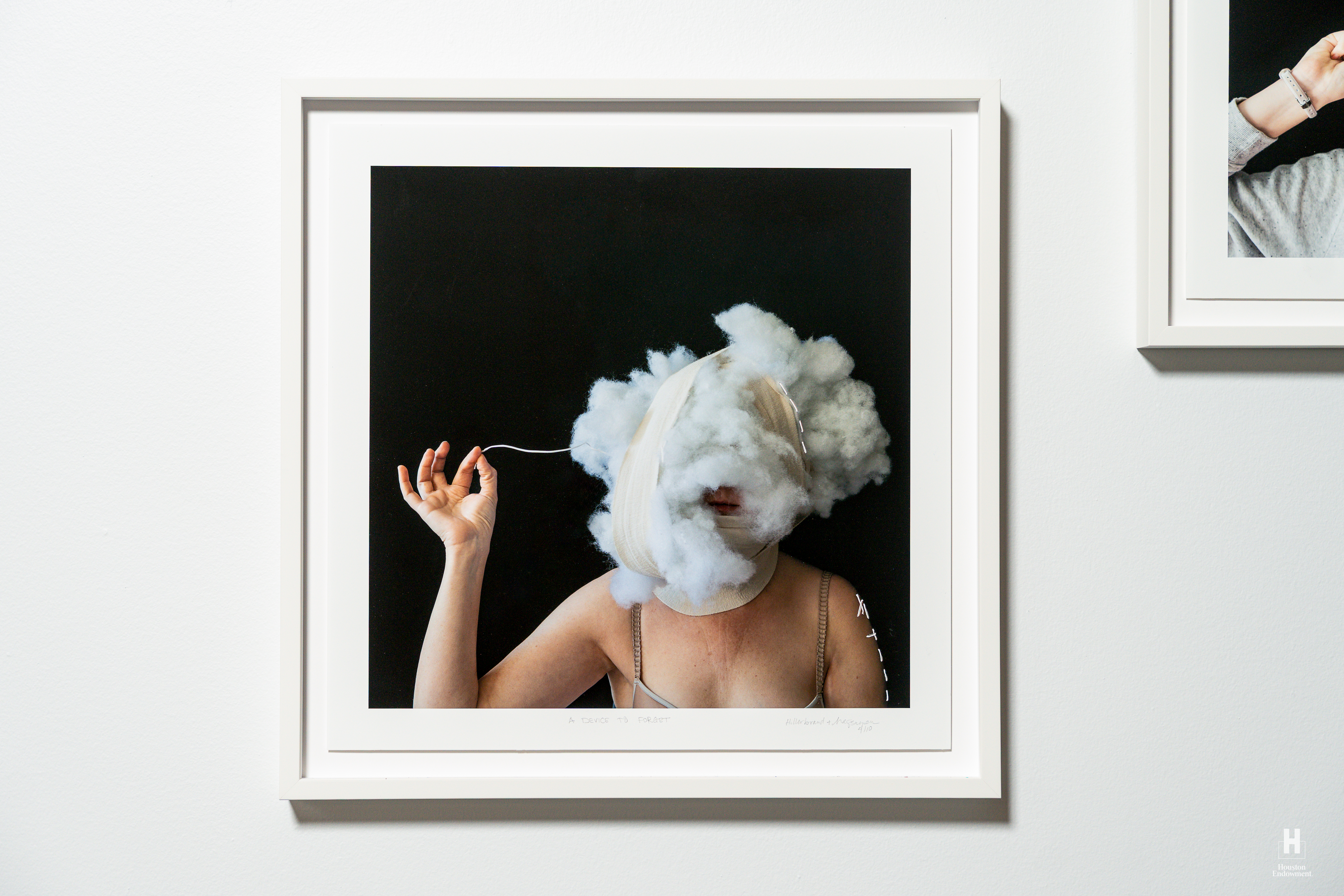 Framed photograph of a bare-shouldered person with bandages and a cotton cloud obscuring their face, tugging a thread.