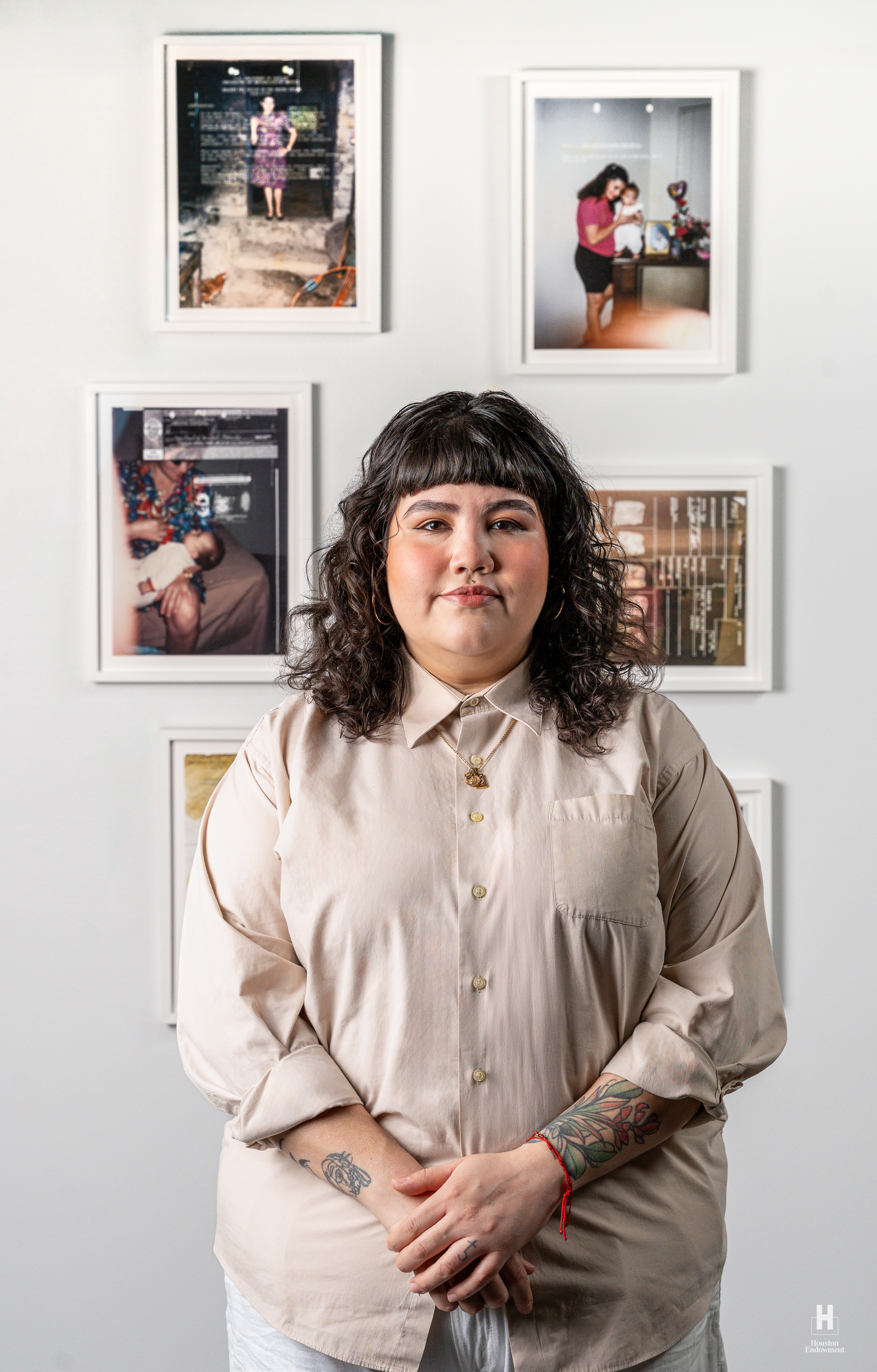 Woman with dark curly hair and bangs wearing a beige shirt, clasping hands to show floral forearm tattoos, in front of 6 framed photos.