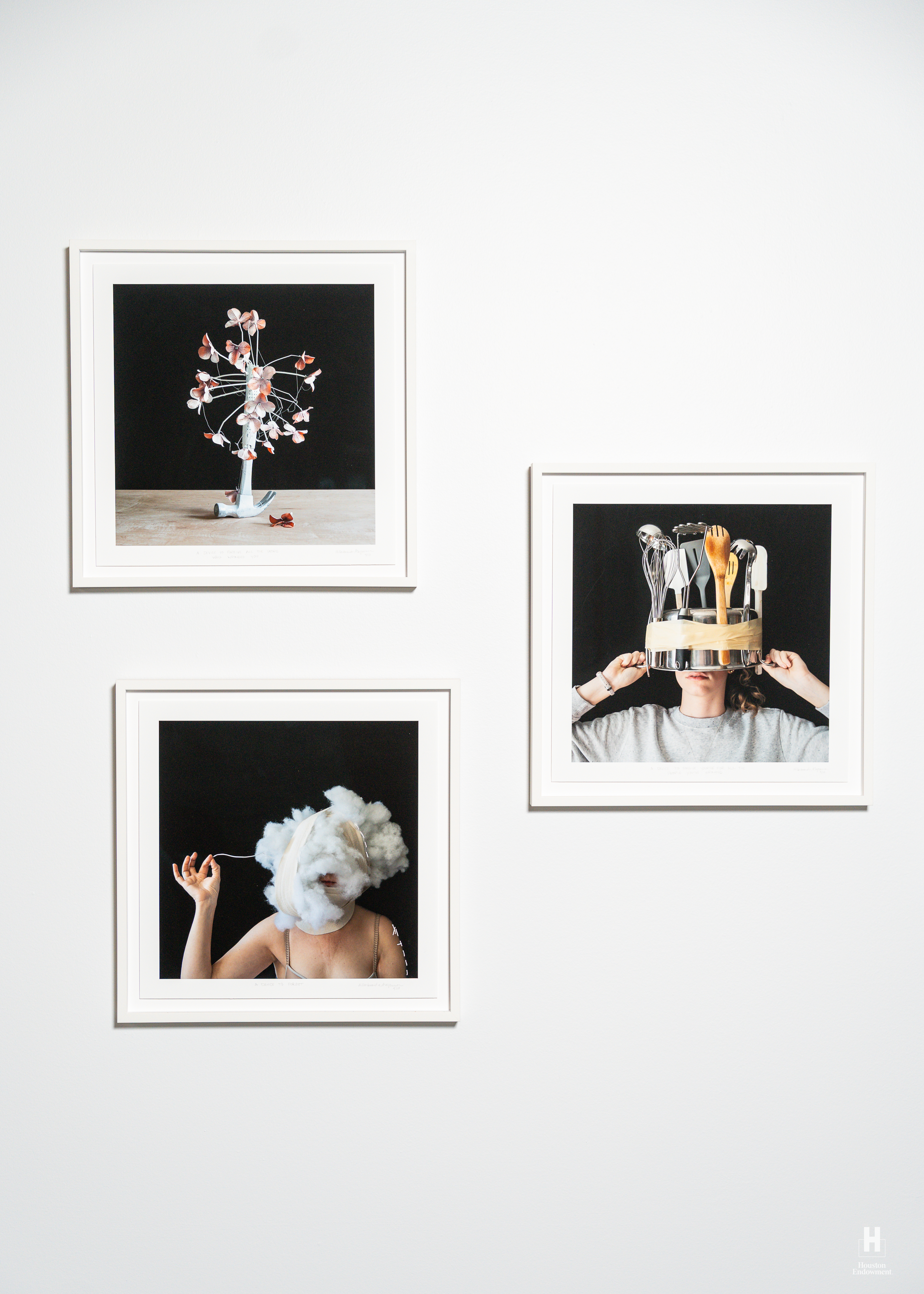 Three framed photos on white wall show a flower sculpture, a person wearing a pot of utensils on head, and a woman with a cotton cloud head.
