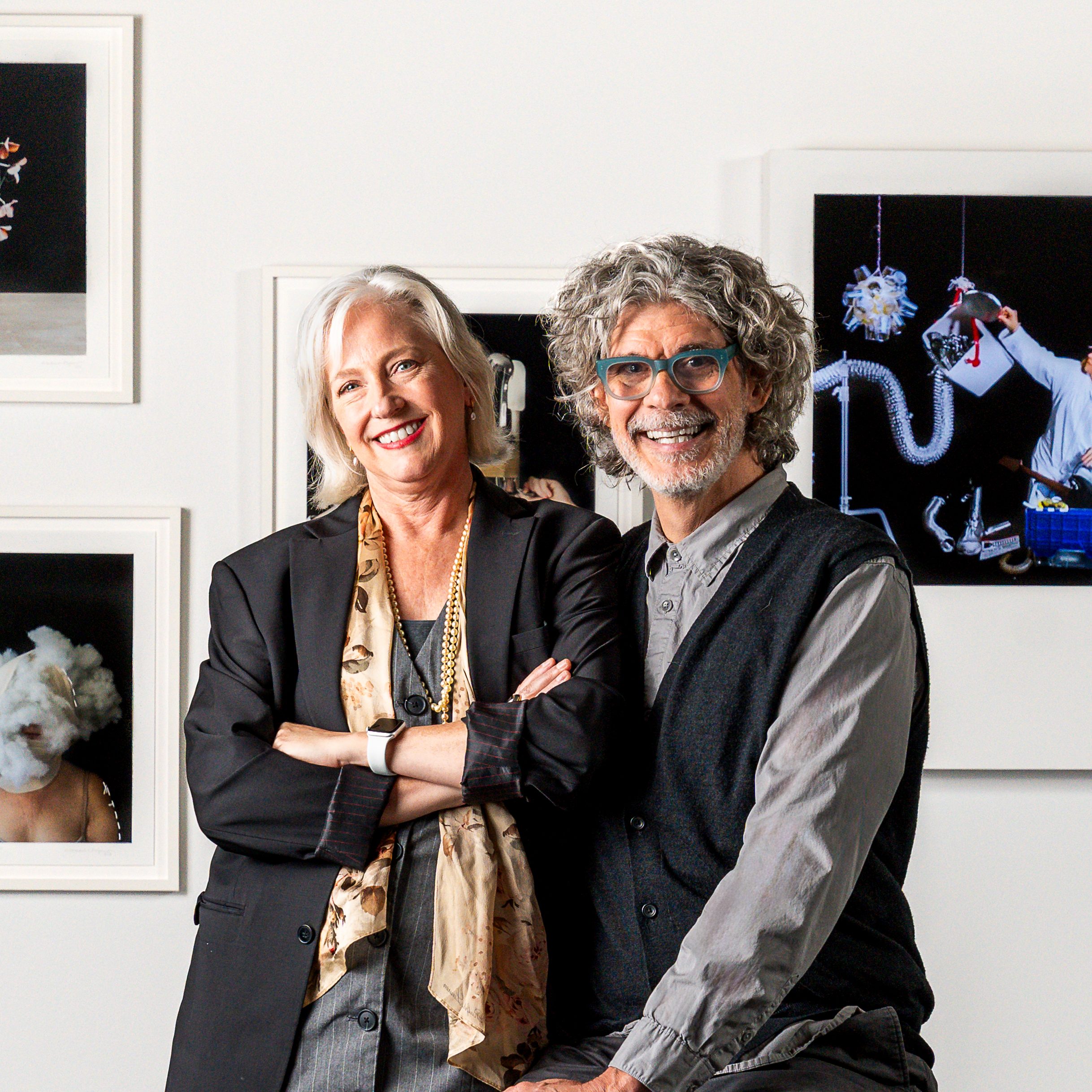 Smiling older woman with white hair in a black blazer and scarf poses with a gray-haired bespectacled man in a vest before framed art.