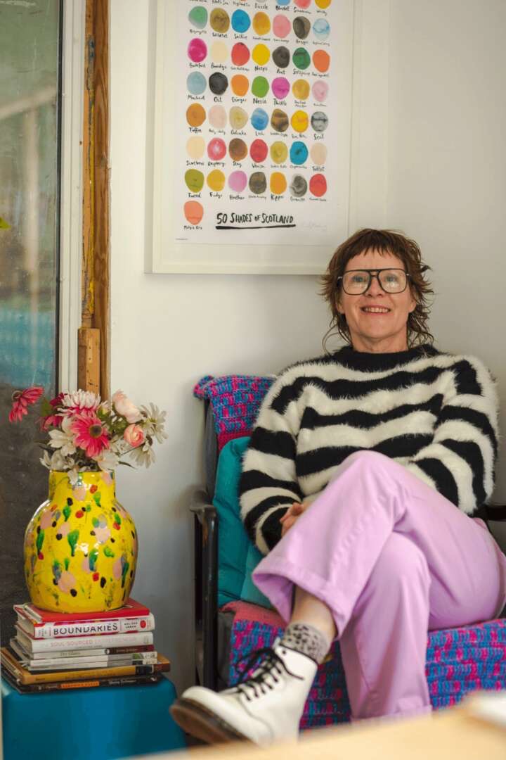 Smiling woman in large glasses and a black-and-white striped fuzzy sweater, lilac pants and white boots seated by a colorful vase.
