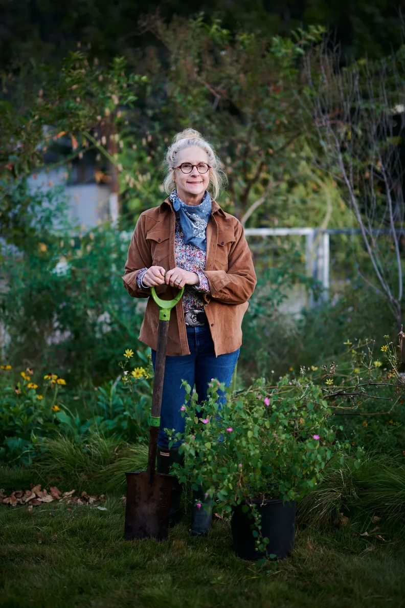 Older woman gardener with glasses and hair in a bun stands holding a green-handled shovel beside a potted shrub sporting small pink flowers.