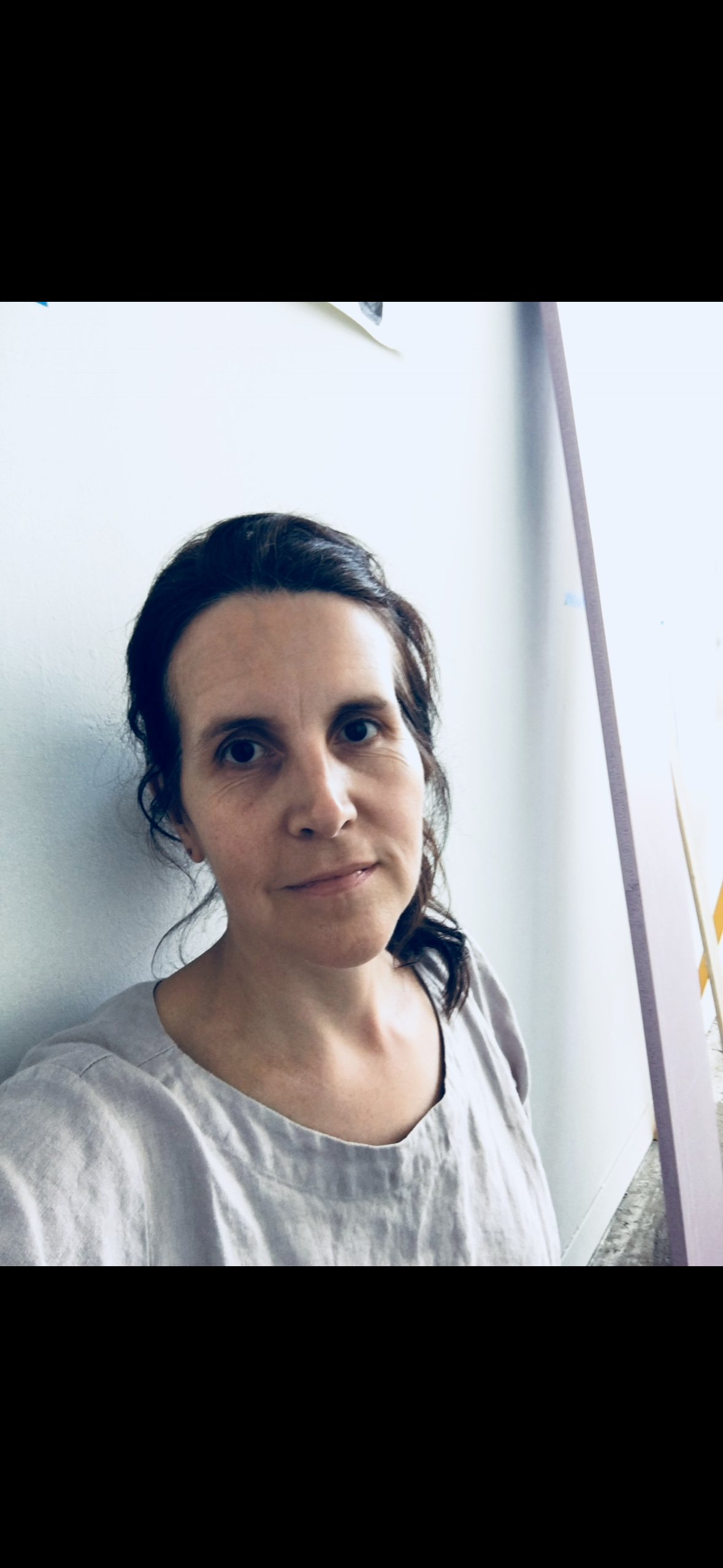 Woman with dark pulled-back hair and soft expression, wearing a pale linen blouse, leaning against a white wall lit by soft window light.