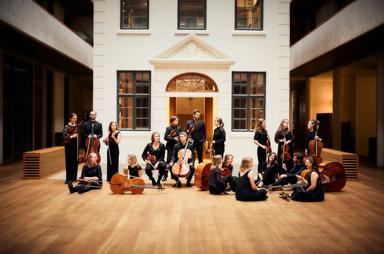 Flere medlemmer av et orkester står i et stort rom foran en hvitmalt bygning. De holder hvert sitt strykeinstrument og er kledd i svart.