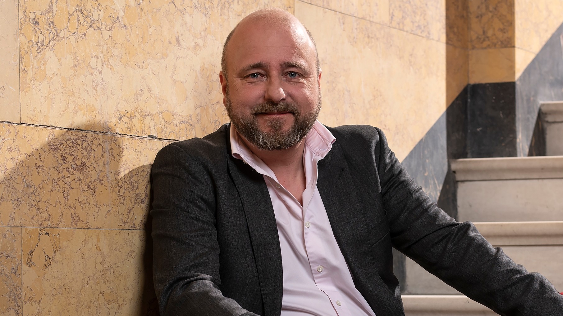 En mann sitter i en trapp iført svart dress og rosa skjorte, og smiler mot kamera.