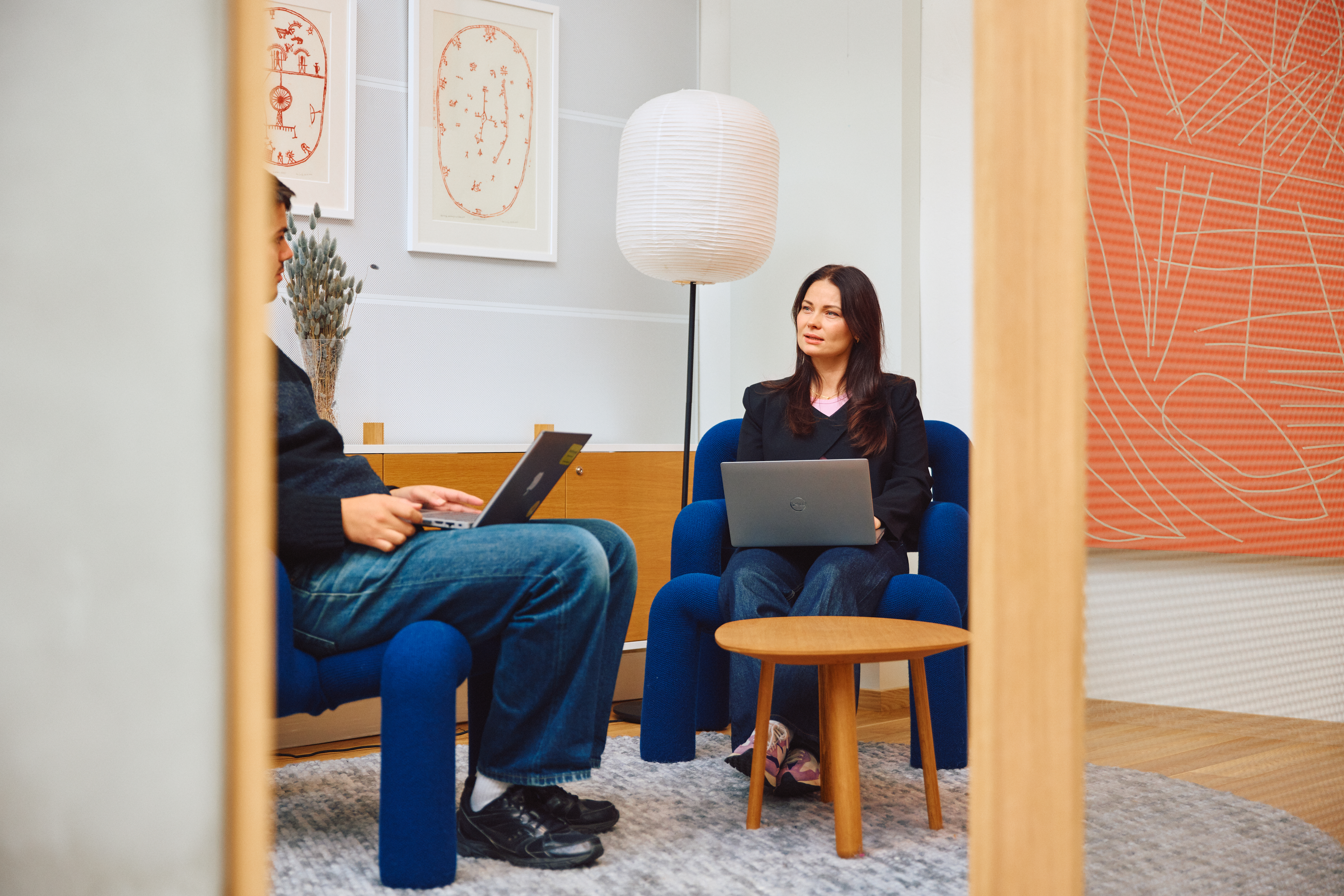 To personer sitter i blå stoler i et moderne og stilrent kontormiljø, med åpne bærbare PC-er på et lite rundt trebord mellom seg. Bakgrunnen inkluderer en hvit gulvlampe i papirdesign, abstrakt kunst på veggen og en stor oransje tekstil med geometrisk mønster. Rommet har minimalistisk innredning med grått teppe og dempet belysning.