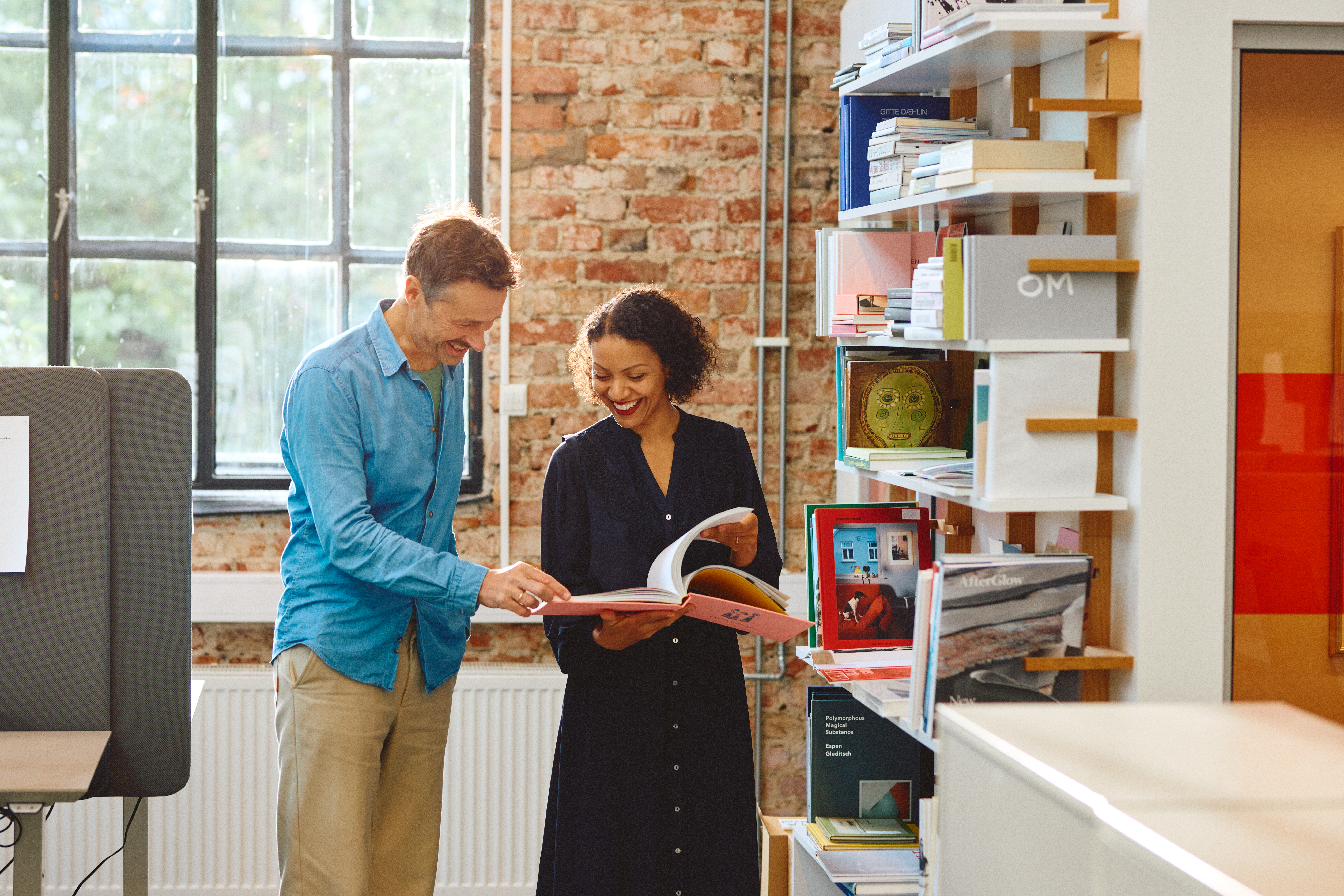 To personer står sammen i et lyst rom og ser i en bok. Den ene peker på noe i boken, mens den andre smiler. Bak dem er en hylle med bøker og en murvegg med et stort vindu.