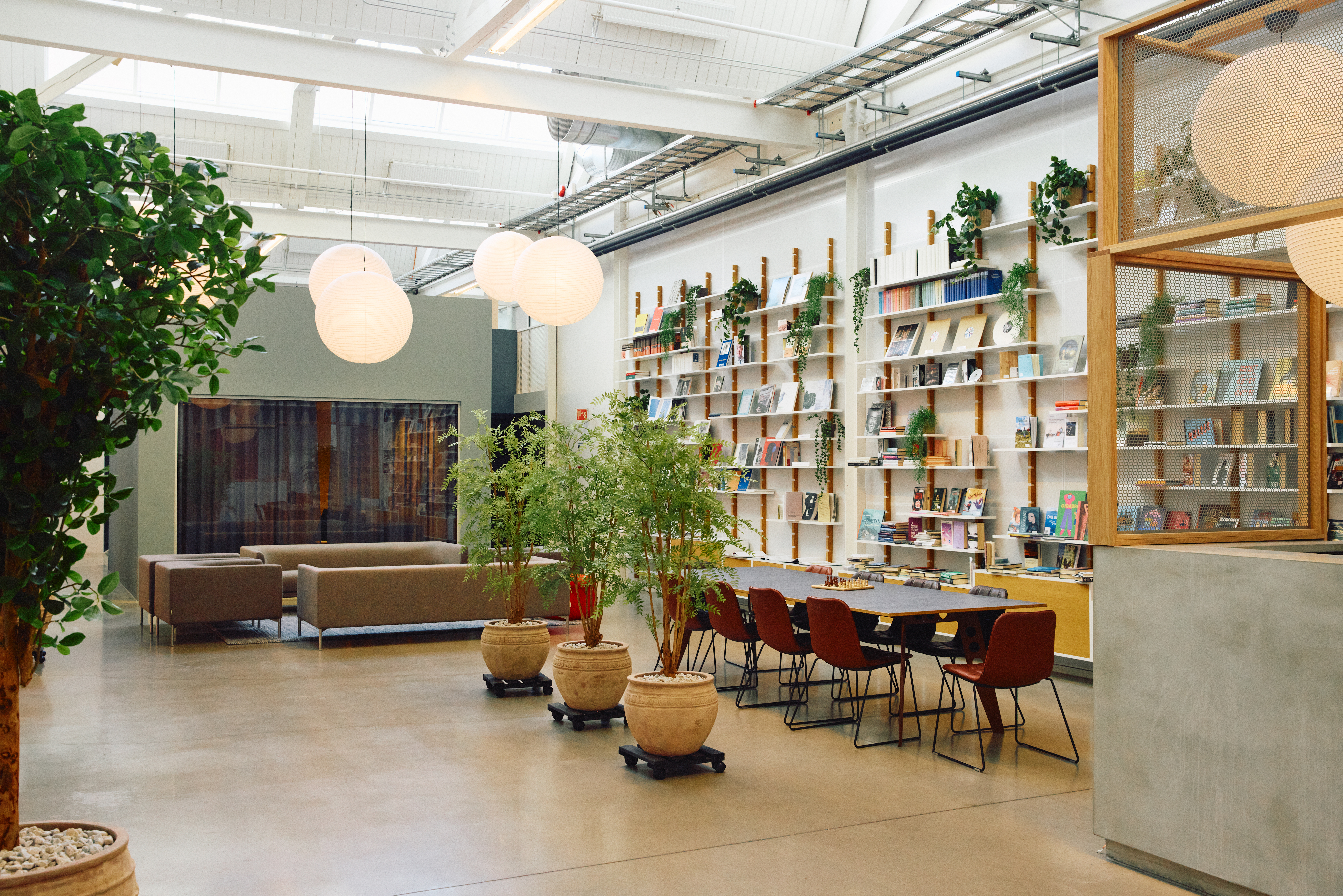 Et lyst og moderne rom med høyt tak, grønne planter i potter, en lang bord med stoler og en vegg med hyller fulle av bøker og planter. Store lamper henger fra taket, og det er en sofagruppe i bakgrunnen.