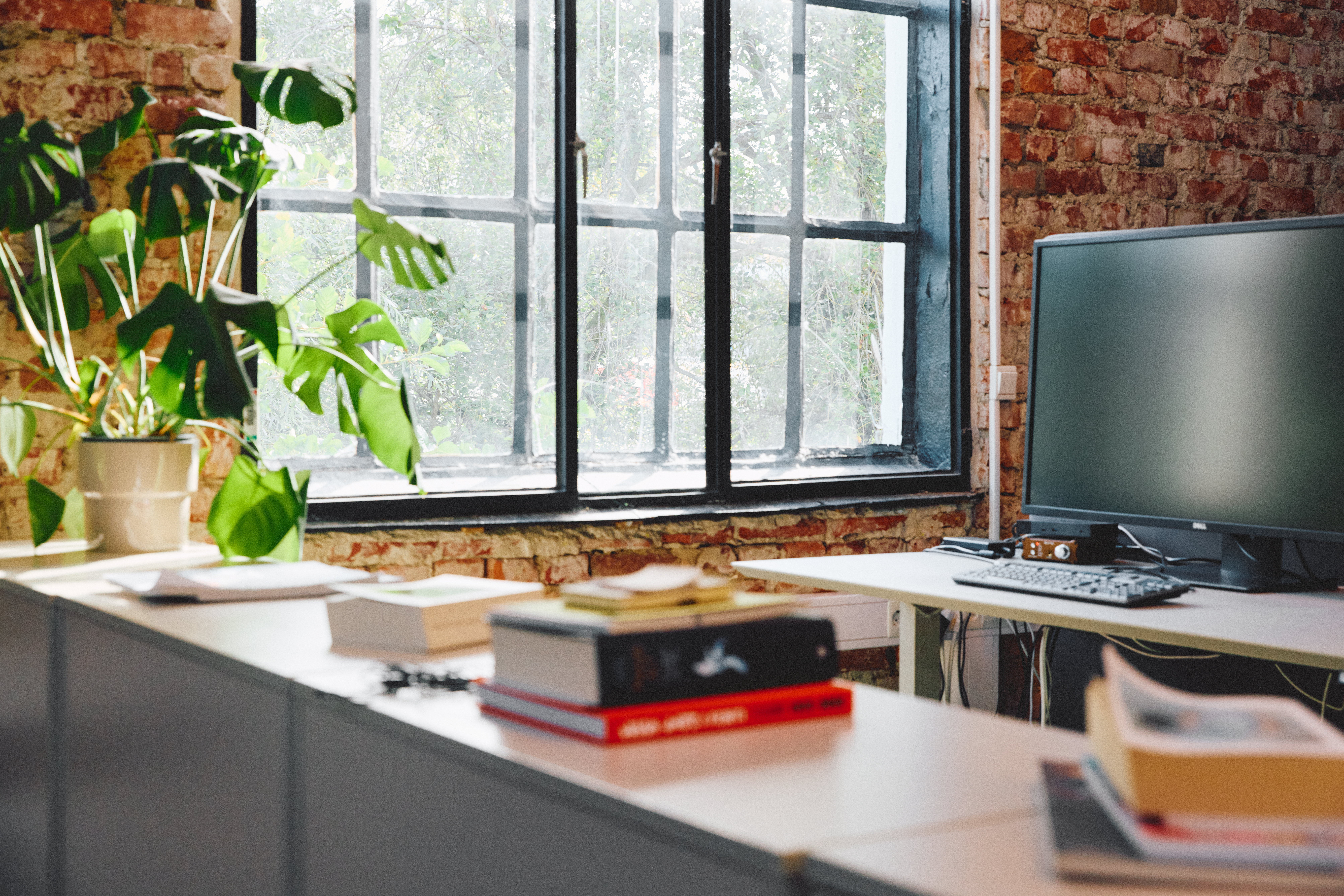 Et kontorlandskap med vindu i bakgrunnen som viser grønne trær. I forgrunnen står en hylle med bøker, og midt i bildet står en skrivepult med en svart PC-skjerm.