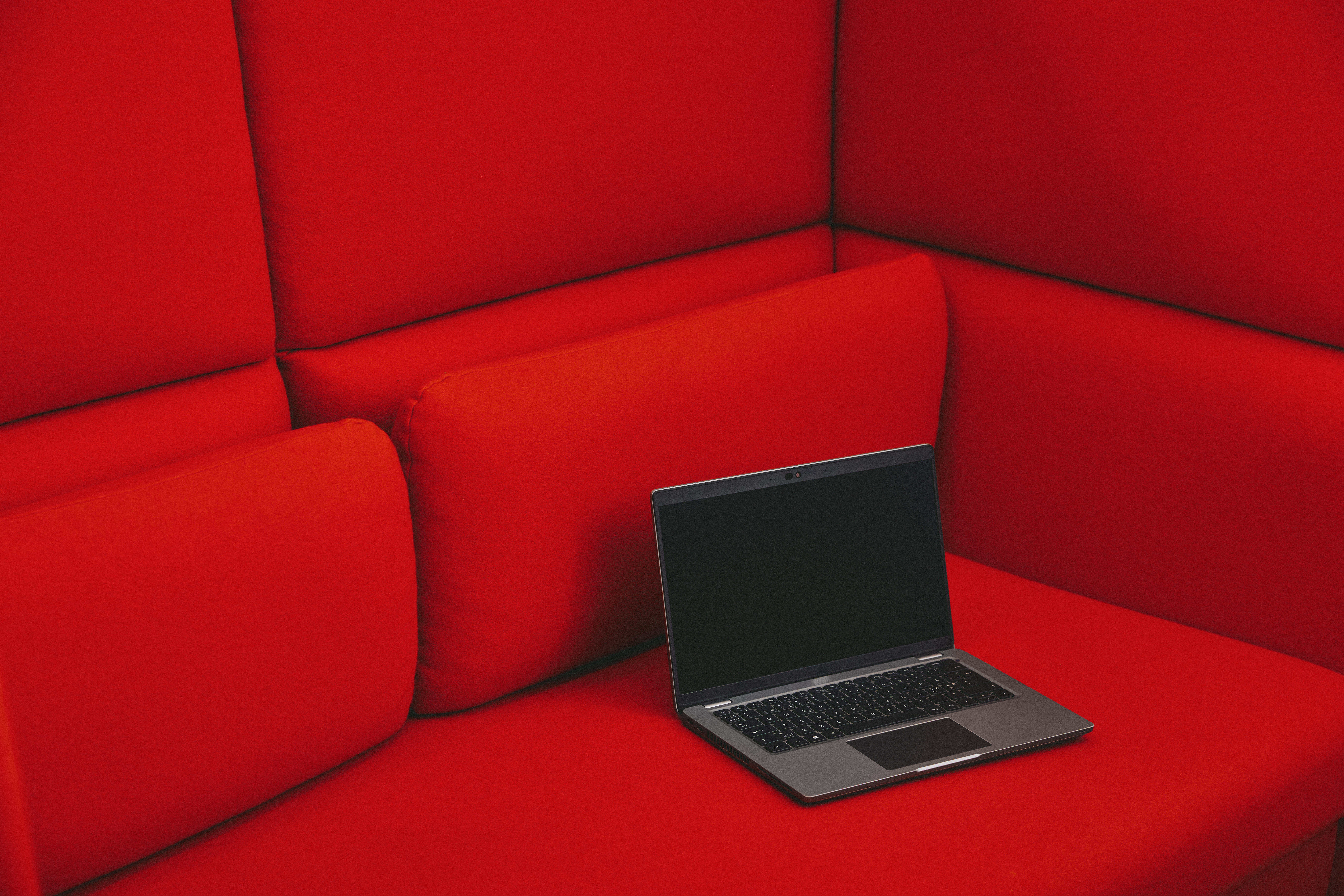 En åpen bærbar PC med svart skjerm står på en rød sofa med høye rygg- og sideputer.