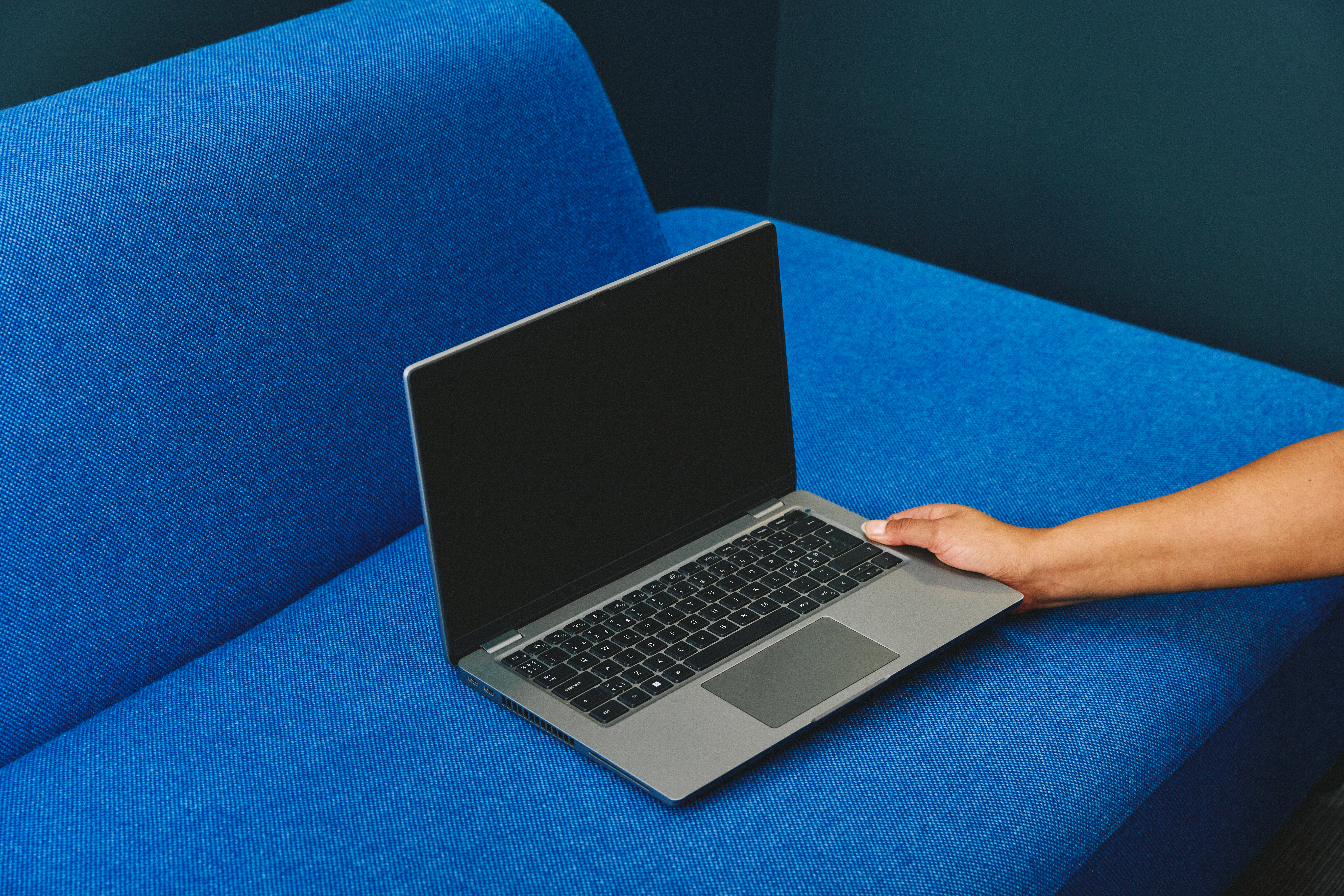 En åpen bærbar PC med svart skjerm står på en blå sofa, mens en hånd hviler på tastaturet.