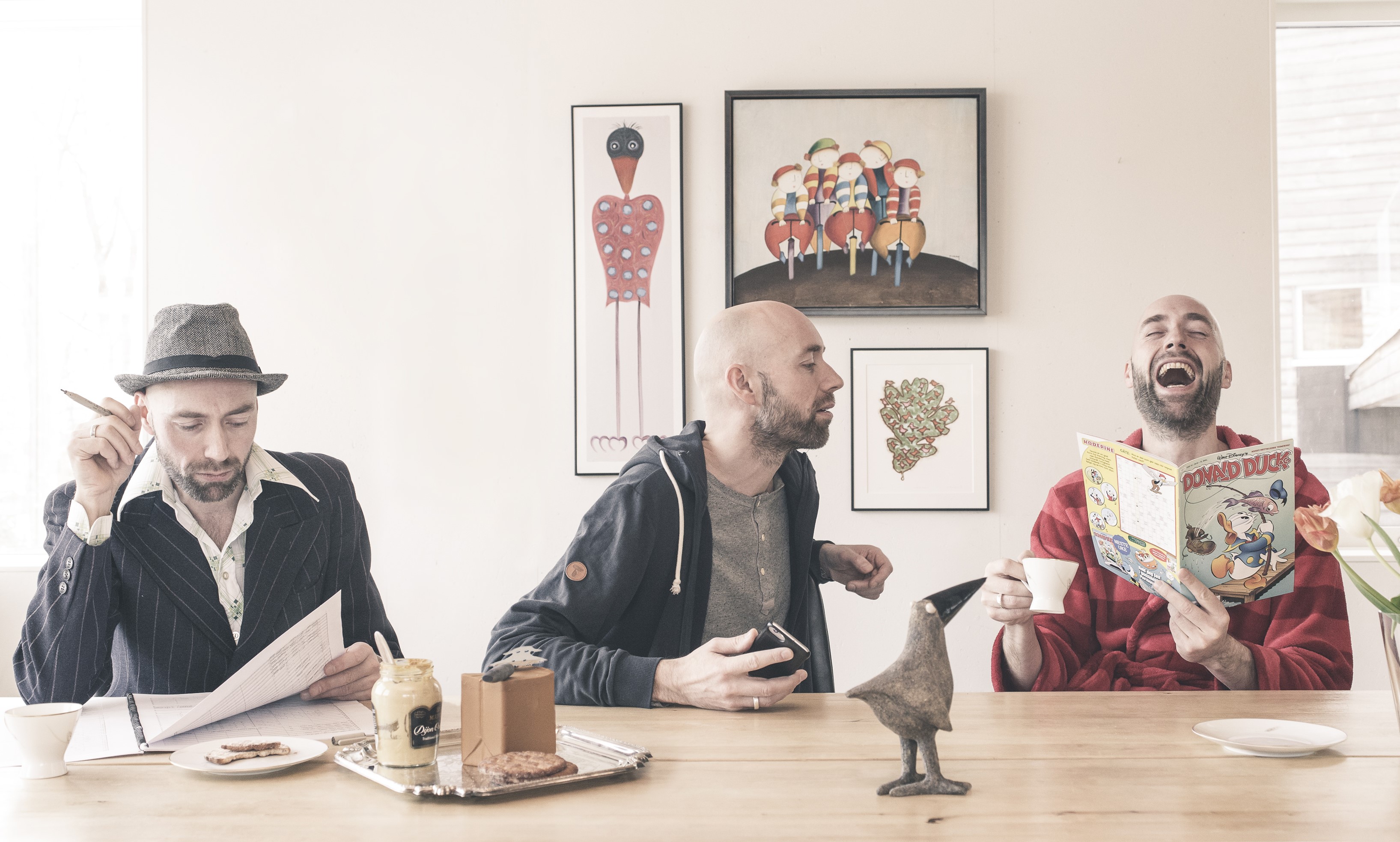 Tre menn sitter ved et trebord og utfører ulike aktiviteter. Den ene, kledd i dress og hatt, studerer papirer med en blyant. Den andre, i hettegenser, snakker eller synger inn i en mikrofon mens han gestikulerer. Den tredje, iført rød morgenkåpe, ler mens han leser et Donald Duck-blad. På bordet står blant annet et brett med peanøttsmør, syltetøy, en kjeks, en kaffekopp og en liten fuglefigur. Bak dem henger fem innrammede kunstverk på veggen. Bildet formidler en humoristisk og kreativ scene med kontrasterende personligheter.
