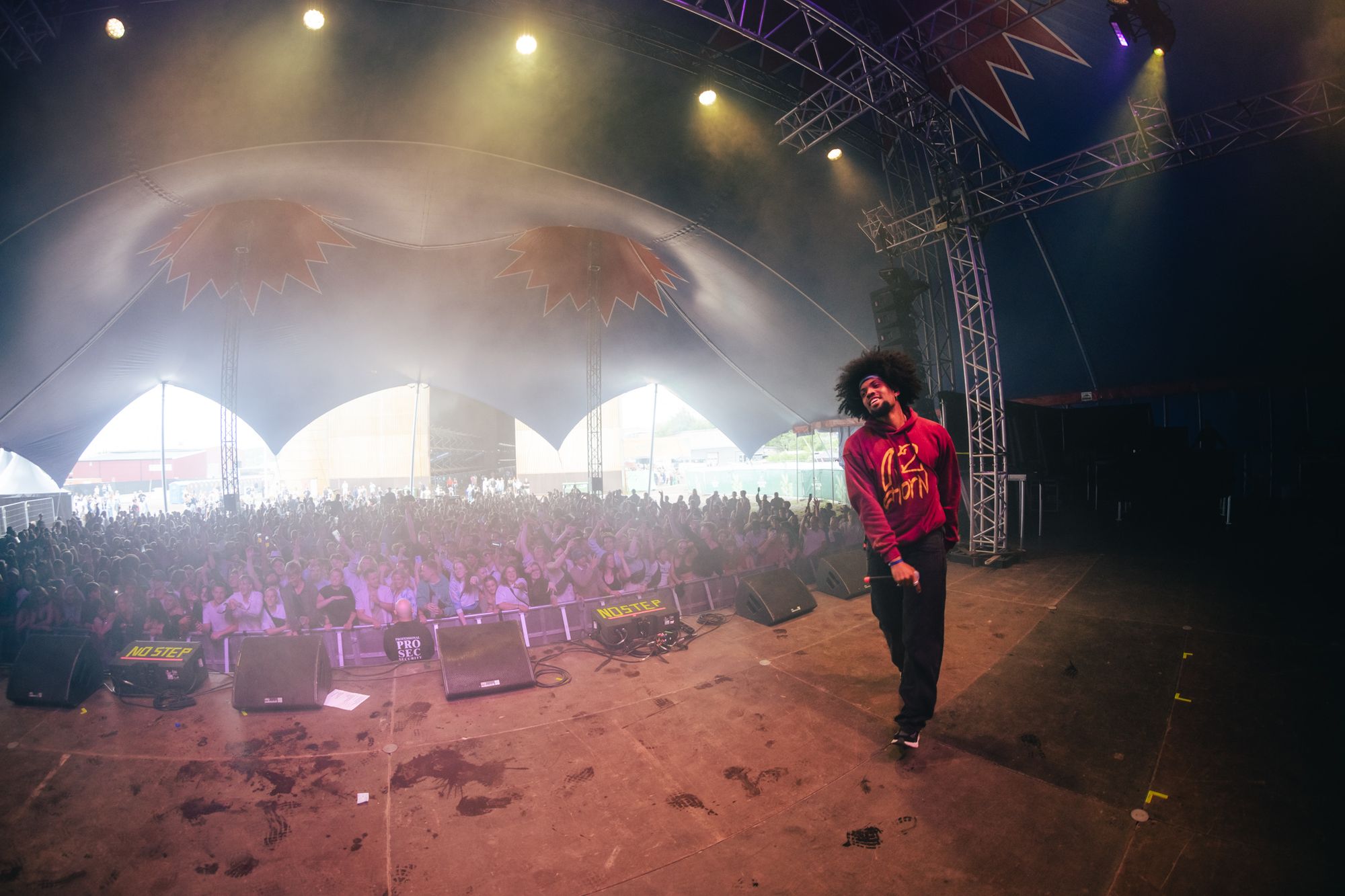 Artist med stort krøllete hår står på en scene inne i et stort telt og holder en mikrofon. Personen har på seg en rød genser med gul tekst og engasjerer publikum, som står tettpakket foran scenen. Scenen er utstyrt med høyttalere og lysrigg, og dekorative elementer henger fra taket. Naturlig lys strømmer inn gjennom buede åpninger i teltstrukturen. Bildet formidler energien og stemningen fra en livekonsert.