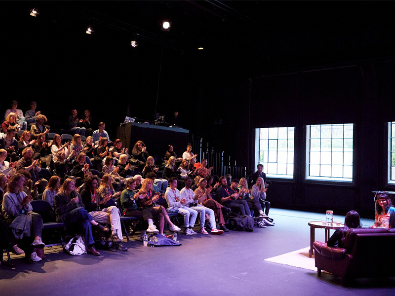 Publikum sitter i amfi-lignende seter i et mørklagt auditorium og applauderer mot en scene der to personer sitter i lenestoler med et lite bord mellom seg. Spotlys lyser opp scenen, mens store vinduer på siden slipper inn naturlig lys. Bildet viser et engasjerende øyeblikk under en samtale eller paneldebatt, med tydelig samspill mellom scene og publikum.