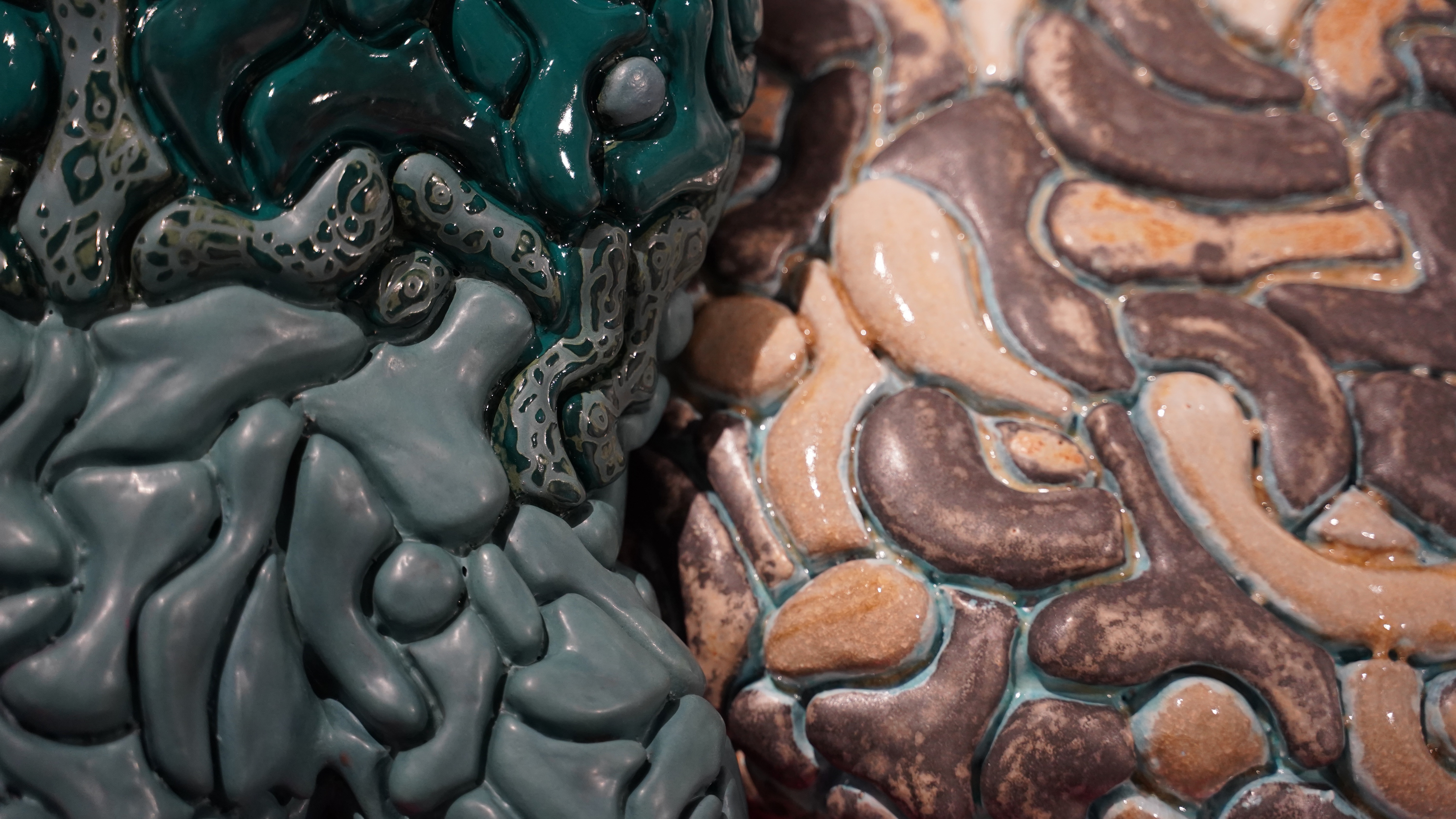 Nærbilde av en keramisk overflate med glaserte, organiske former i grønne, beige og brune toner, satt sammen i et mønster som minner om celler eller steinformer.