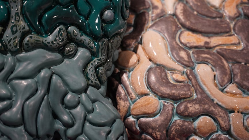 Nærbilde av en keramisk overflate med glaserte, organiske former i grønne, beige og brune toner, satt sammen i et mønster som minner om celler eller steinformer.