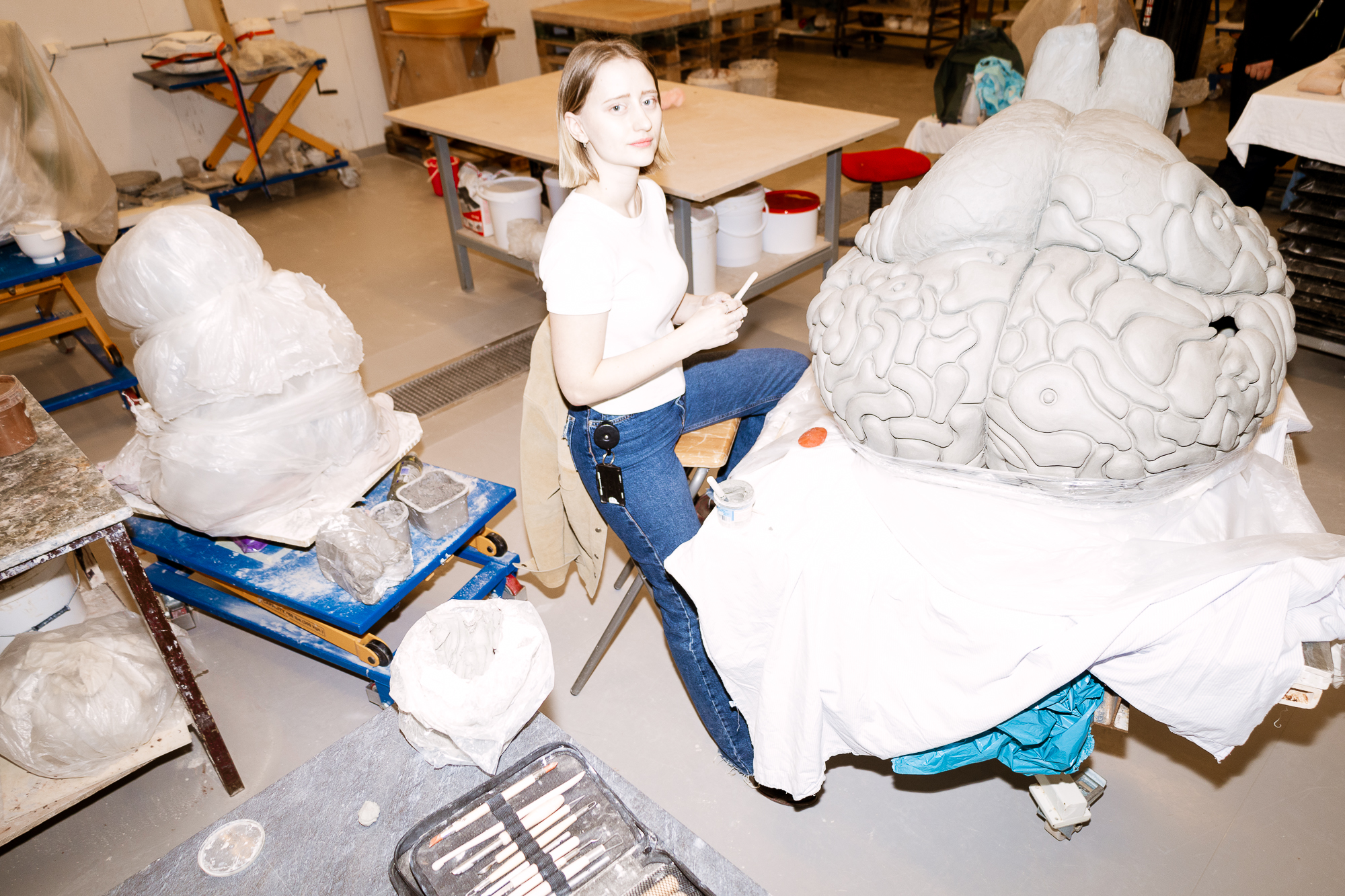 En kunstner sitter i et verksted og arbeider med en stor skulptur av en hjerne, laget i et hvitt materiale som gips eller leire. Verkstedet er fylt med ulike verktøy og materialer som brukes i skulpturprosessen.