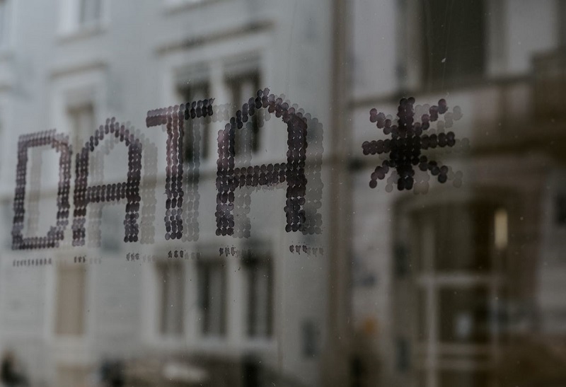 Glassvindu med ordet 'DATA*' skrevet med små sirkulære prikker. I bakgrunnen skimtes en uskarp bygning, noe som tyder på at vinduet tilhører et kontor- eller næringslokale. Typografien og ordvalget antyder en tilknytning til teknologi, dataanalyse eller digital innovasjon.