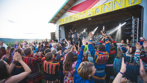 Utendørs konsert eller festival med stor folkemengde foran en scene merket 'MARKOMEANNU'. Mange i publikum er kledd i tradisjonelle samiske klær og klapper eller løfter hendene i feiring. På scenen opptrer artister under sterke scenelys, og stemningen er livlig og feststemt. Bildet formidler et øyeblikk av felles glede og kulturelt fellesskap, trolig under en samisk kulturfestival.