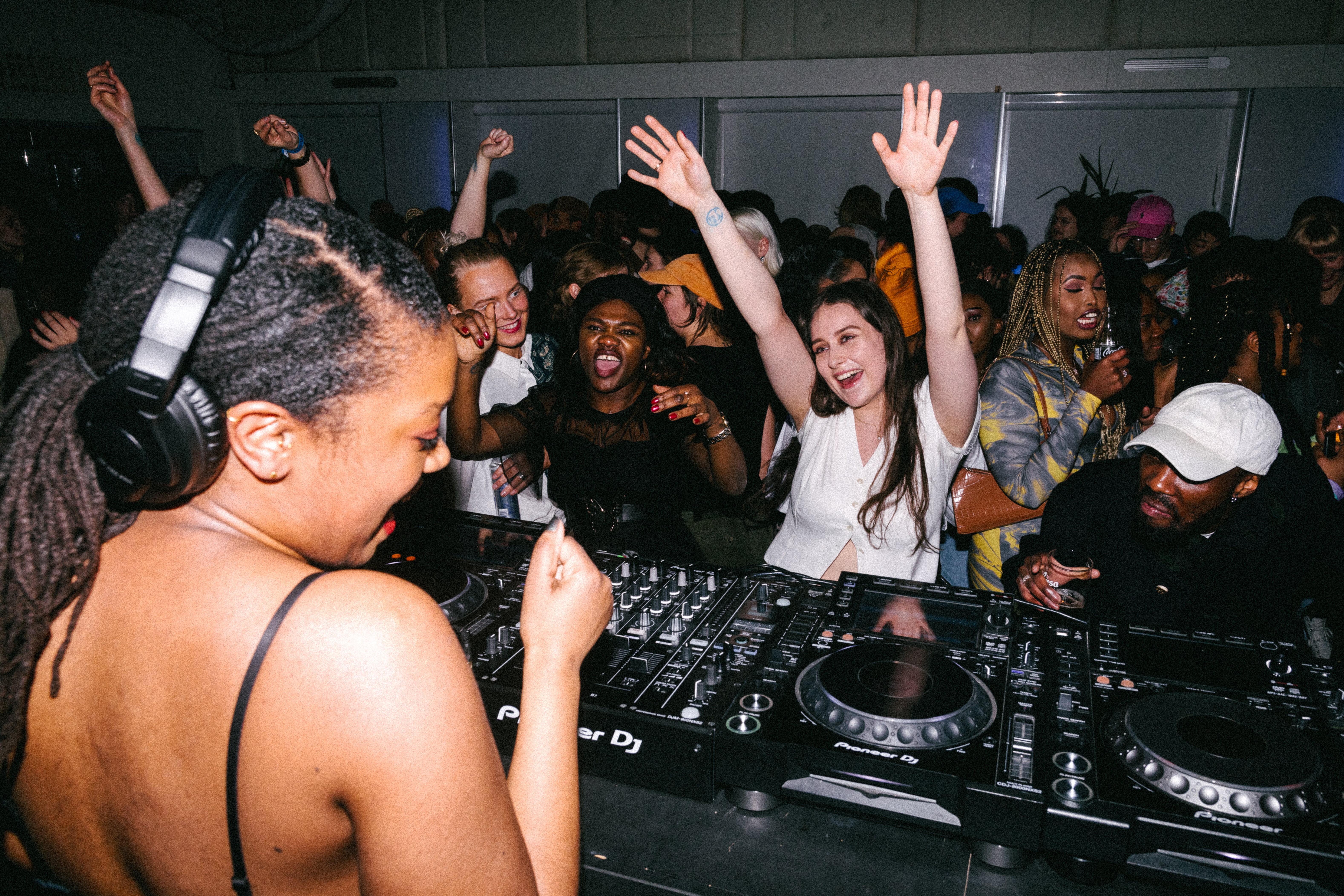 DJ med hodetelefoner spiller musikk på en Pioneer DJ-kontroller foran en dansende folkemengde. Personen er kledd i svart og står i forgrunnen, mens publikum i bakgrunnen smiler, hever hendene og beveger seg til musikken. Fargerik belysning og tett stemning gir bildet en energisk og festlig atmosfære, typisk for en klubb eller musikkarrangement.