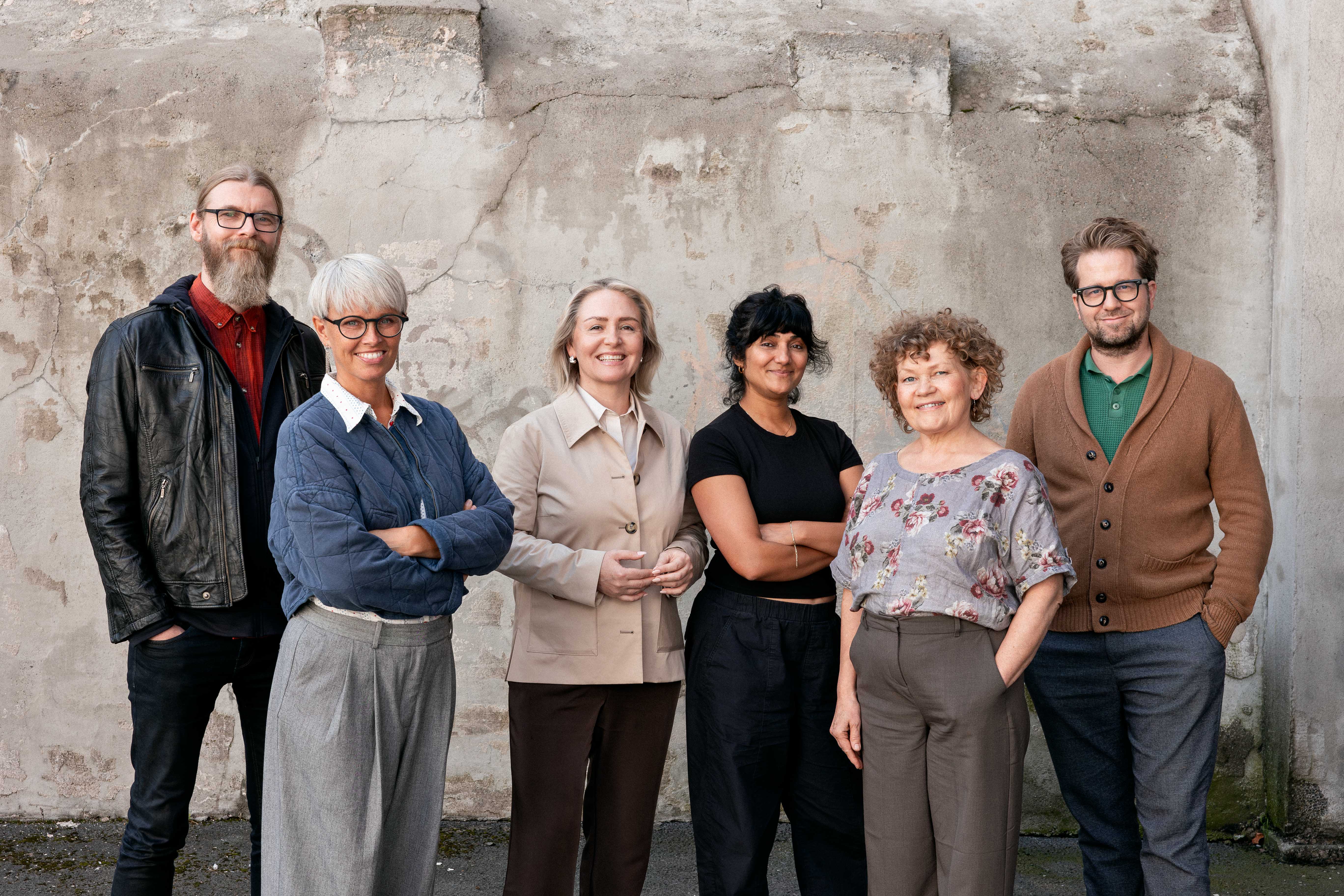 Seks voksne personer står på rekke foran en slitt betongvegg utendørs. De er kledd i en blanding av uformelle og semi-formelle klær, som skjorter, cardigans og bukser. Gruppen viser variasjon i stil og fremtoning, og poserer som for et gruppebilde. Bildet formidler fellesskap og individualitet, og kan representere et team, en sosial gruppe eller et lokalt fellesskap.